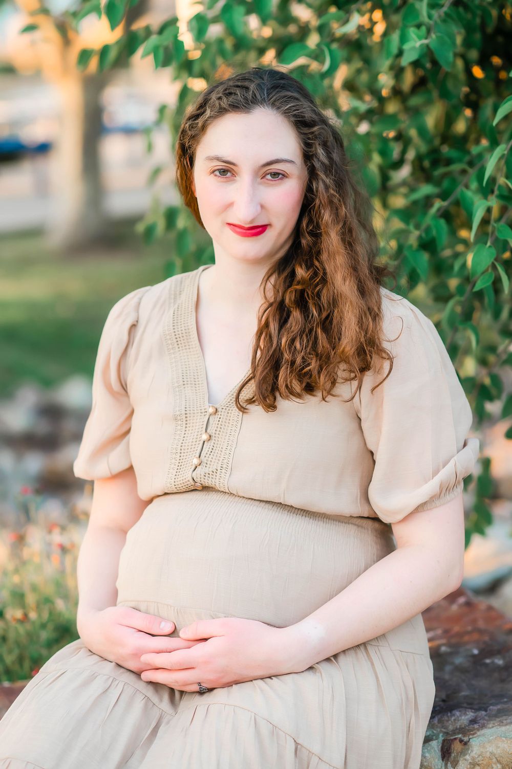 Pregnant woman in beige dress cradling baby bump while sitting outdoors under green foliage.