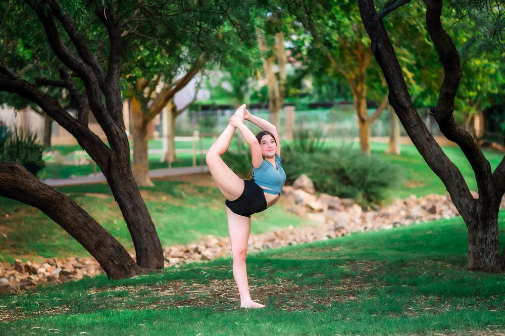Female dancer balancing in Lord of the Dance yoga pose on green grass between trees.