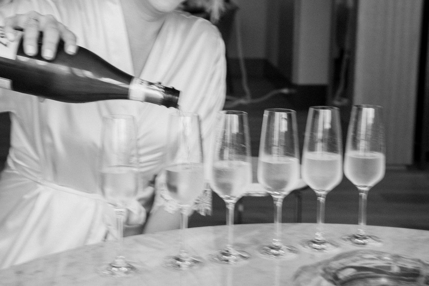 Black and white photo of a woman in a robe pouring champagne into a row of elegant flute glasses on a table.