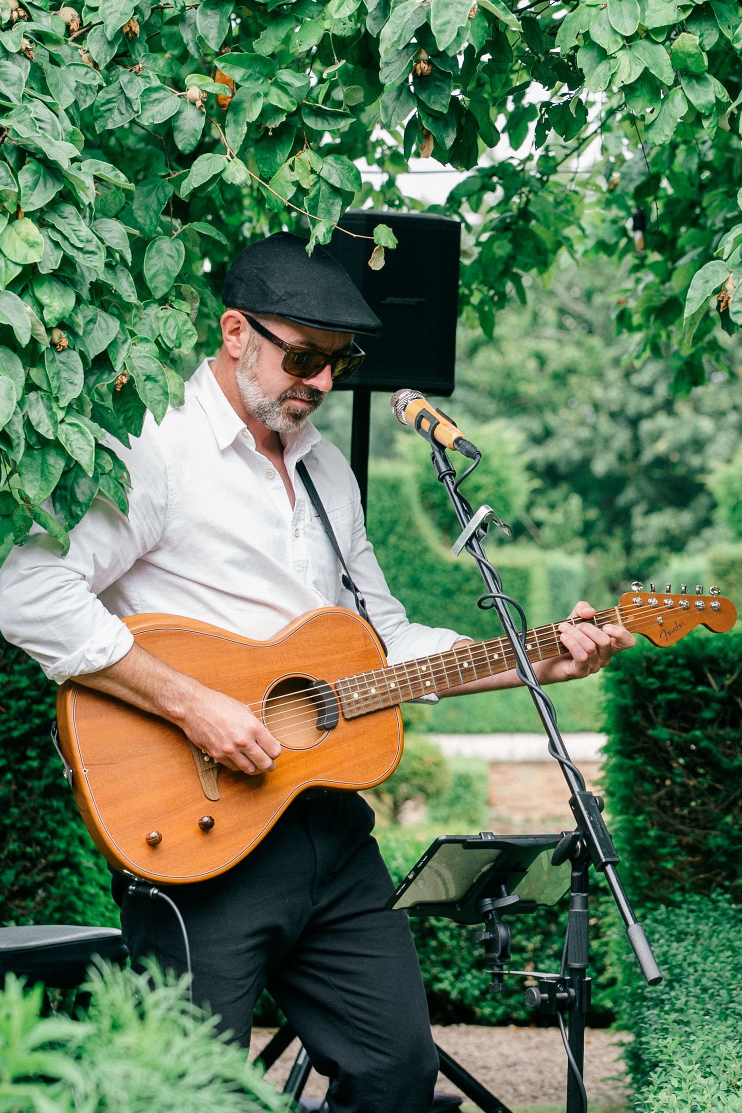 Middle-aged male guitarist in black hat and sunglasses playing acoustic guitar outdoors near microphone stand.