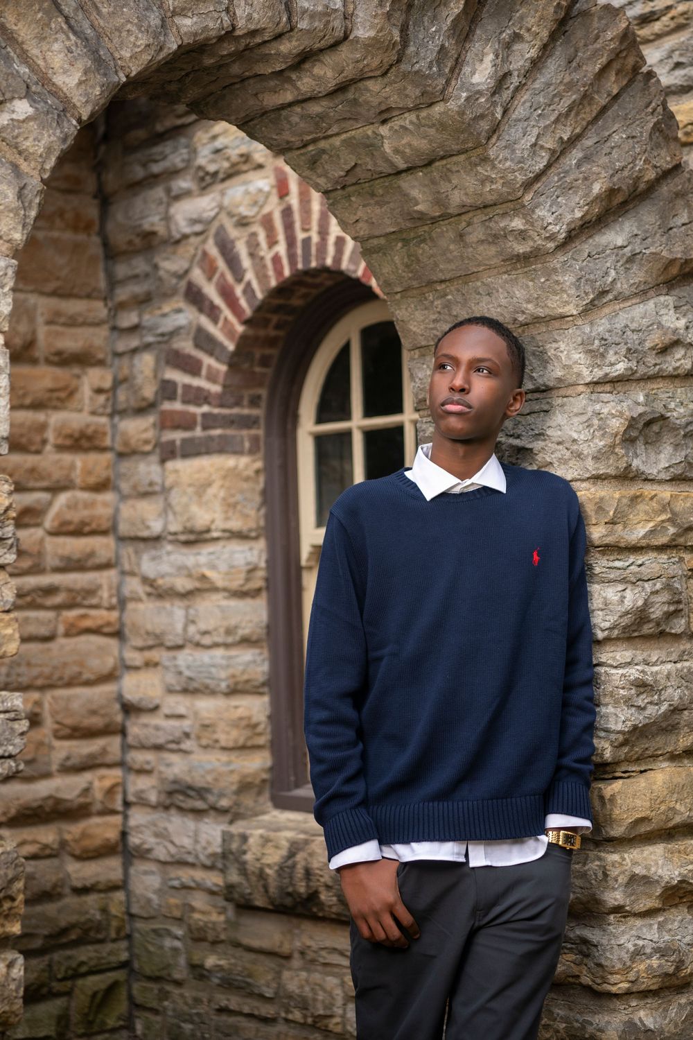 Young man in navy Polo Ralph Lauren sweater leaning against stone arch wall, gazing upward thoughtfully.
