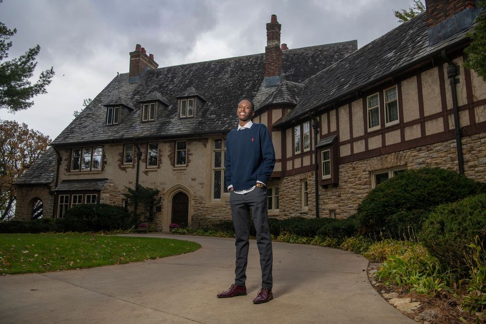 Man standing on curved driveway before a large Tudor-style stone mansion surrounded by autumn trees.