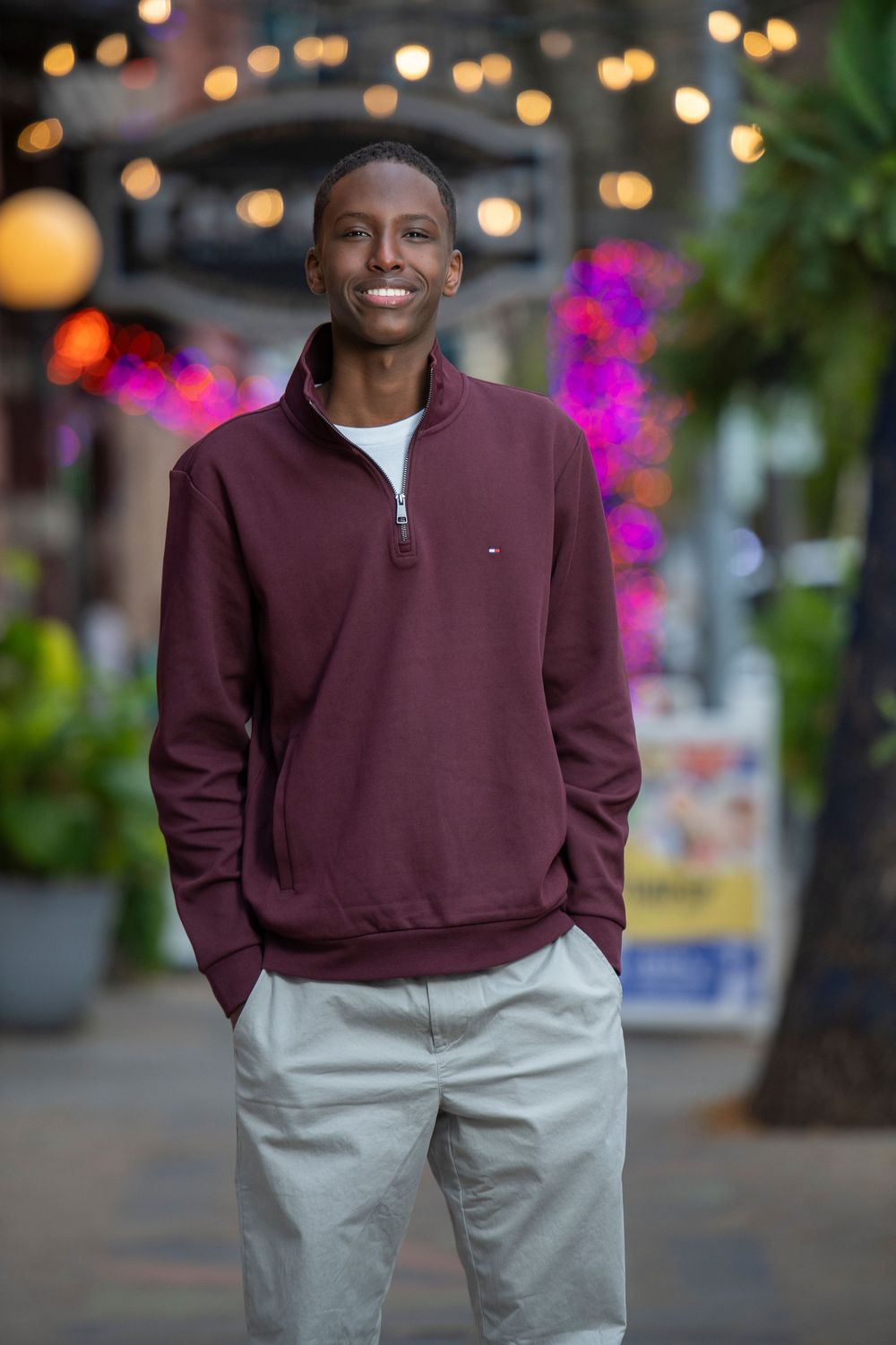 Smiling young man in maroon quarter-zip sweatshirt and khaki pants standing outdoors with bokeh lights.