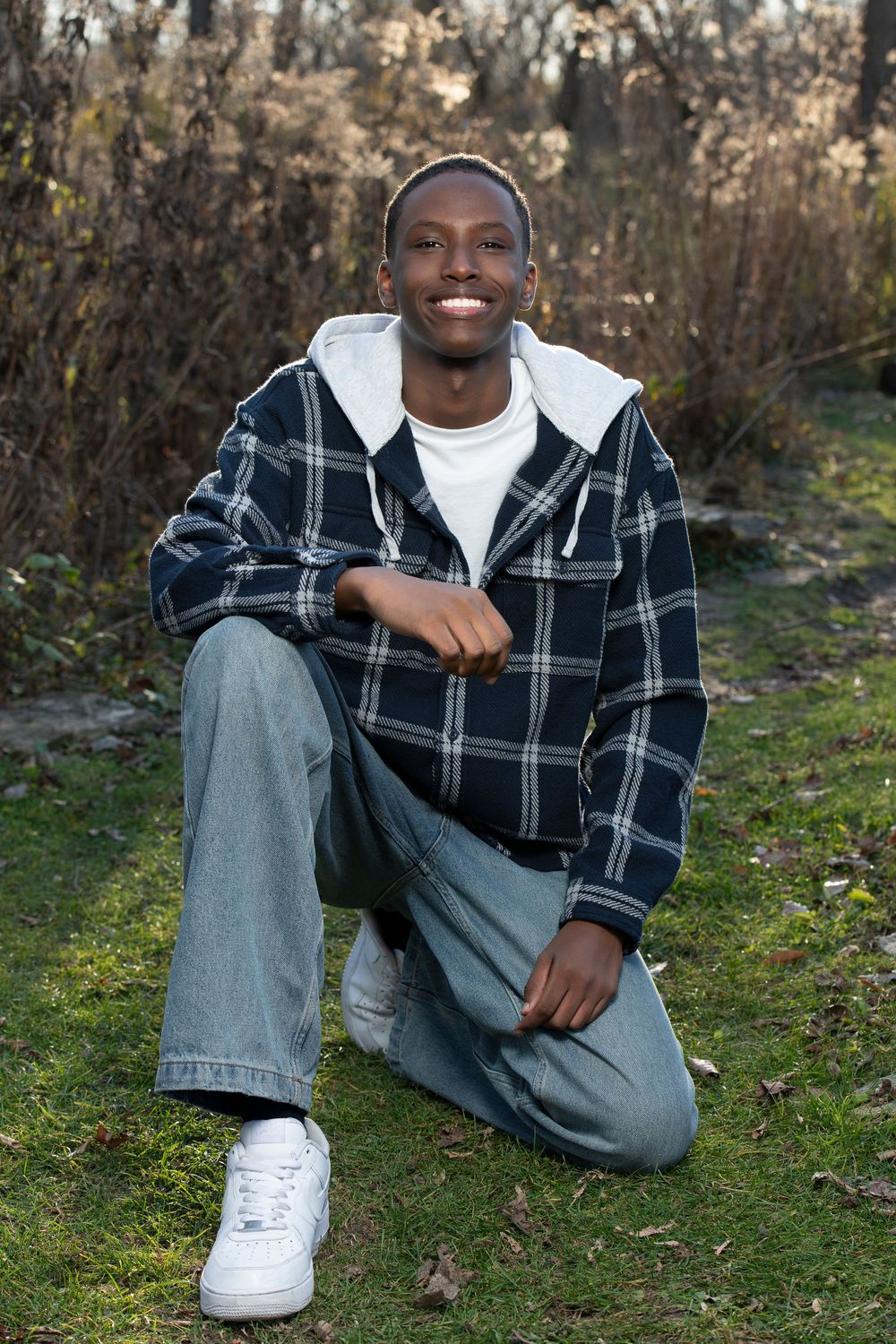 Smiling young Black man in plaid hoodie jacket and jeans sitting outdoors on grass with natural foliage background.