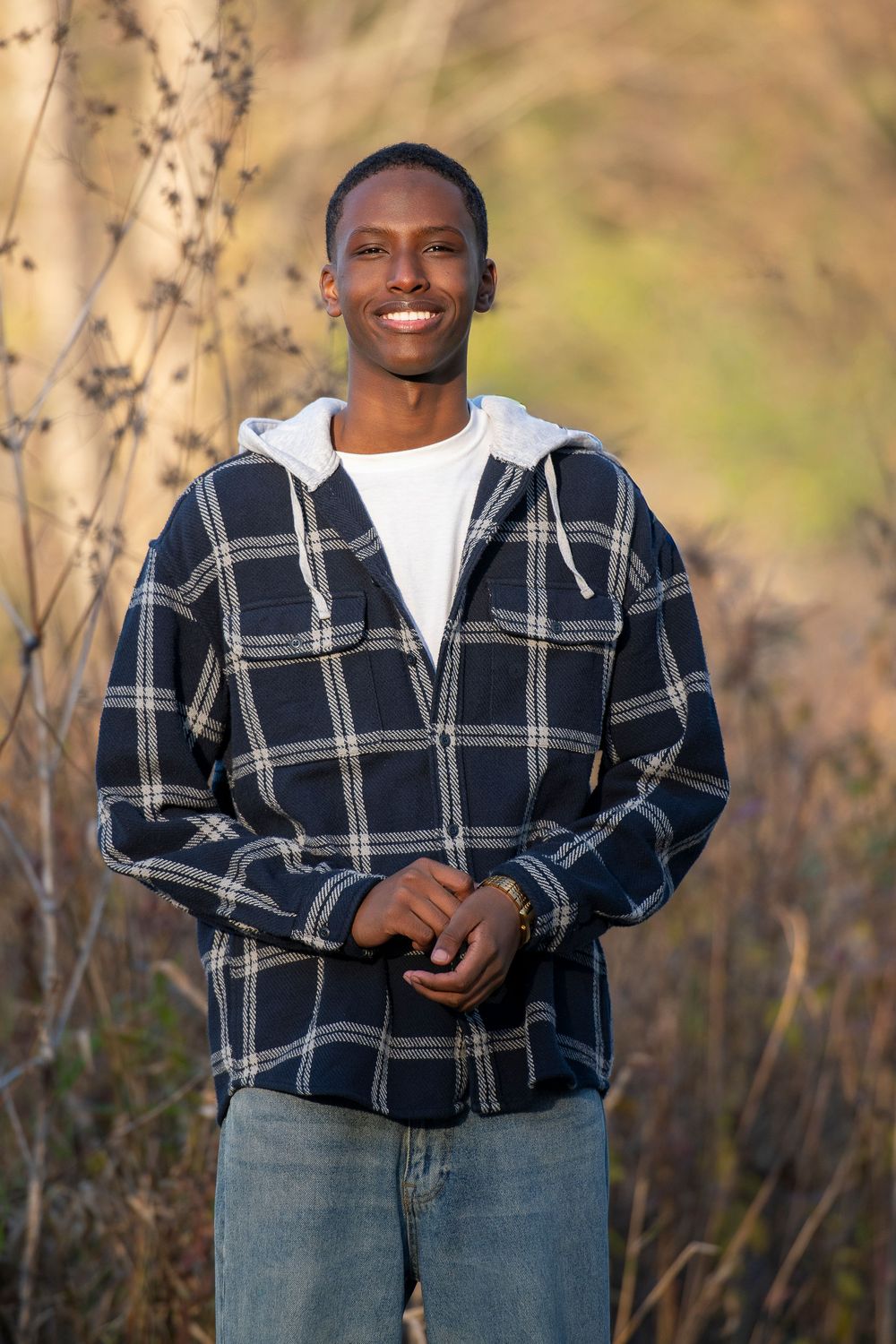 Young Black man smiling outdoors, wearing plaid hooded jacket and jeans, standing among dry branches in natural light.