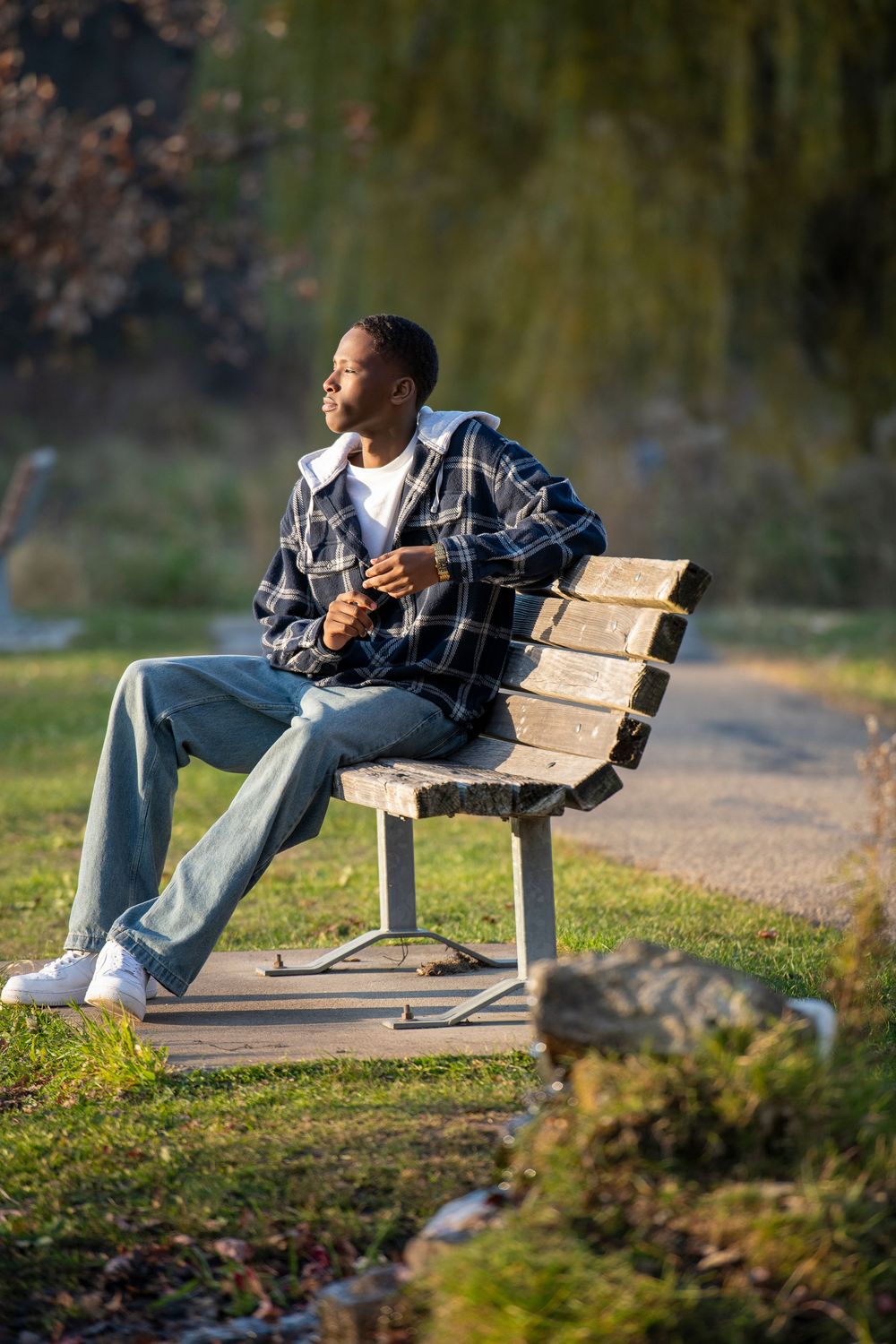 Young Black man in plaid jacket and jeans sitting relaxed on a wooden park bench enjoying sunny day outdoors.