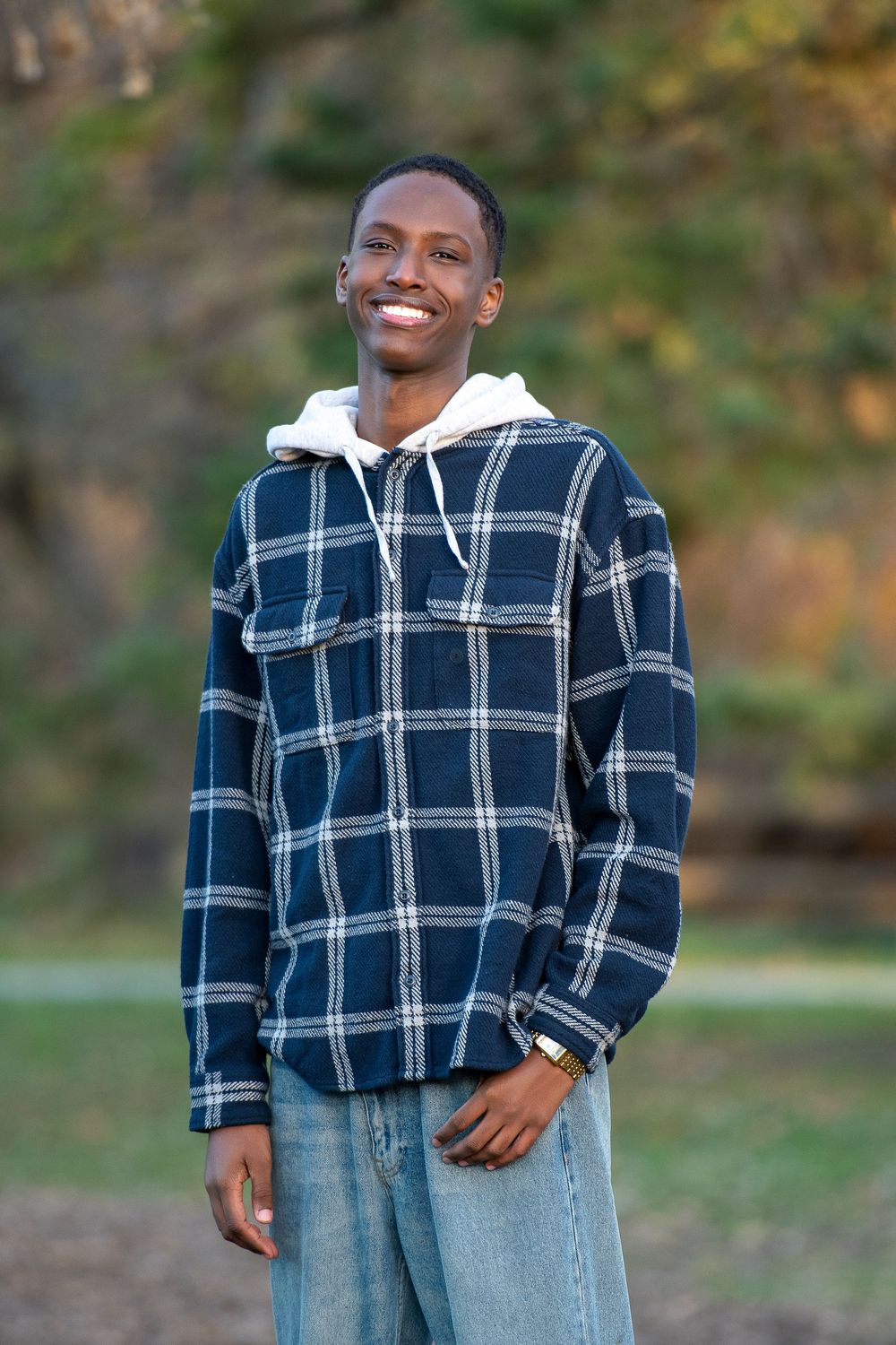 Smiling young Black man in plaid flannel shirt over white hoodie and jeans posing outdoors in a park.