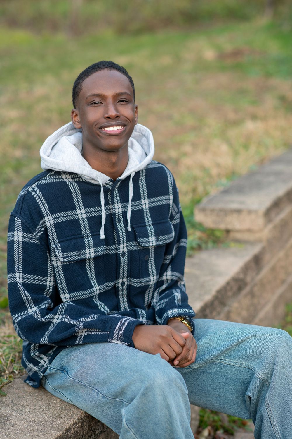 Smiling young Black man in plaid hoodie and jeans sitting on stone steps outdoors in a casual portrait.