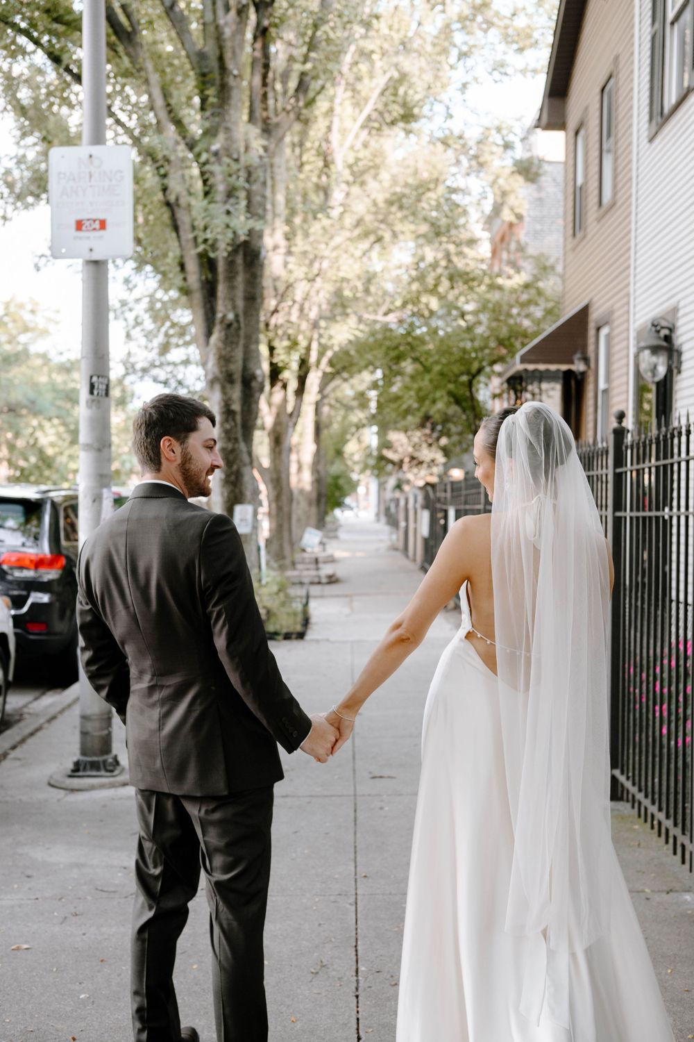 A romantic wedding couple walks down a tree-lined city sidewalk holding hands.