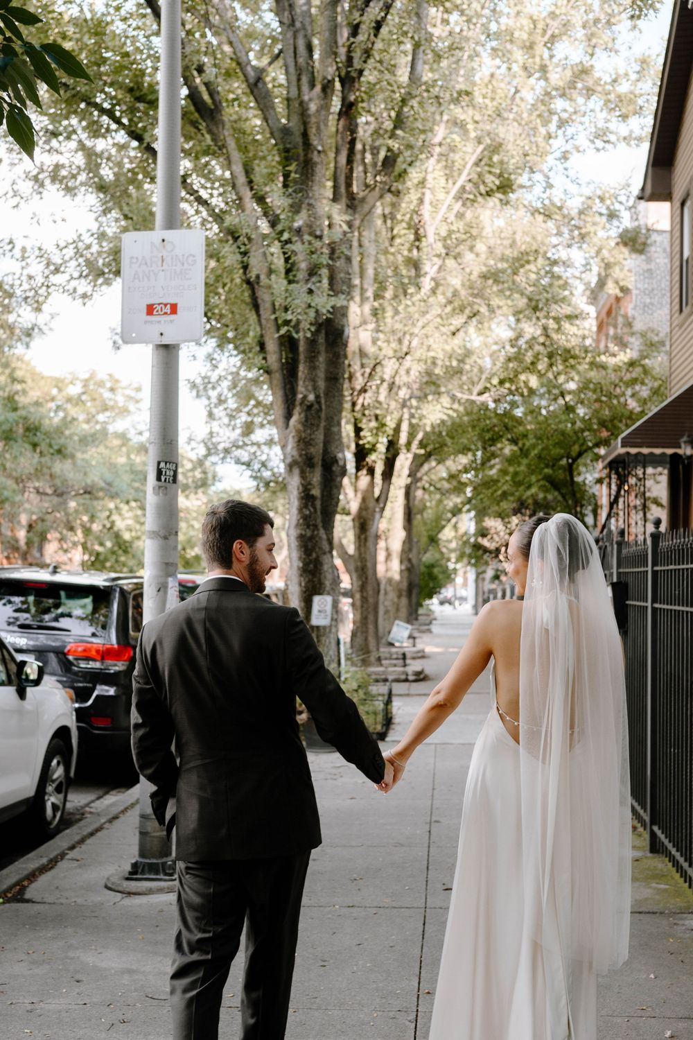 A couple walking down a tree-lined sidewalk, with the bride in a flowing white dress and veil.