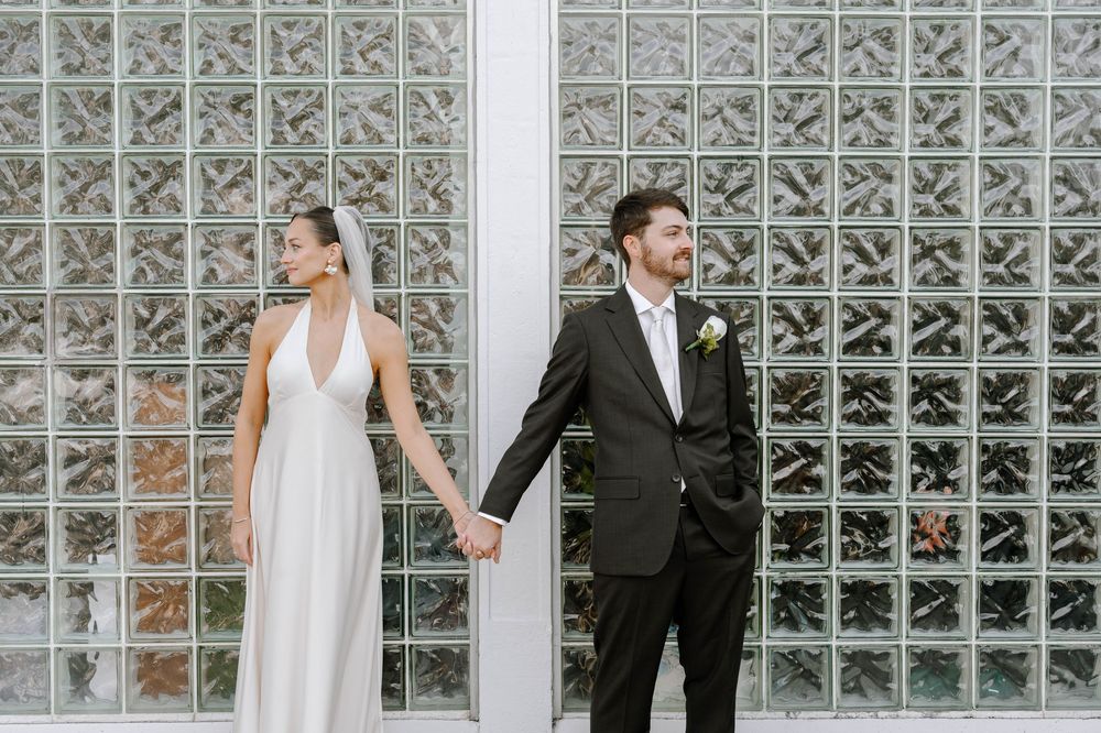 Newlyweds pose together against a glass block wall in an urban setting.