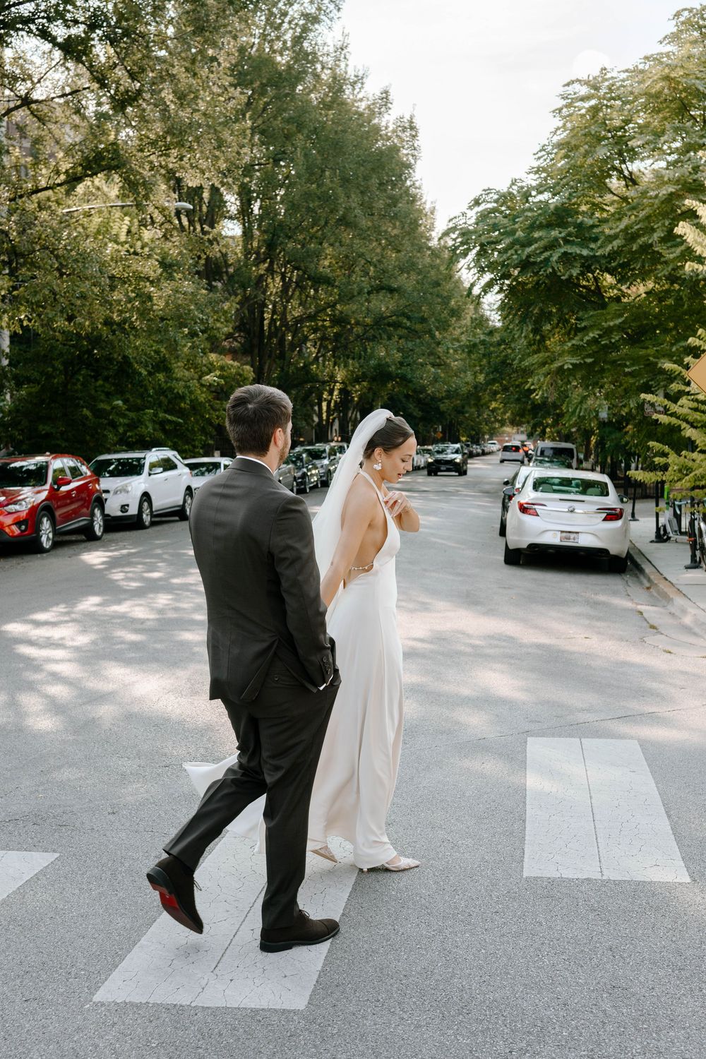 A playful wedding couple dances in a parking lot surrounded by trees and cars.