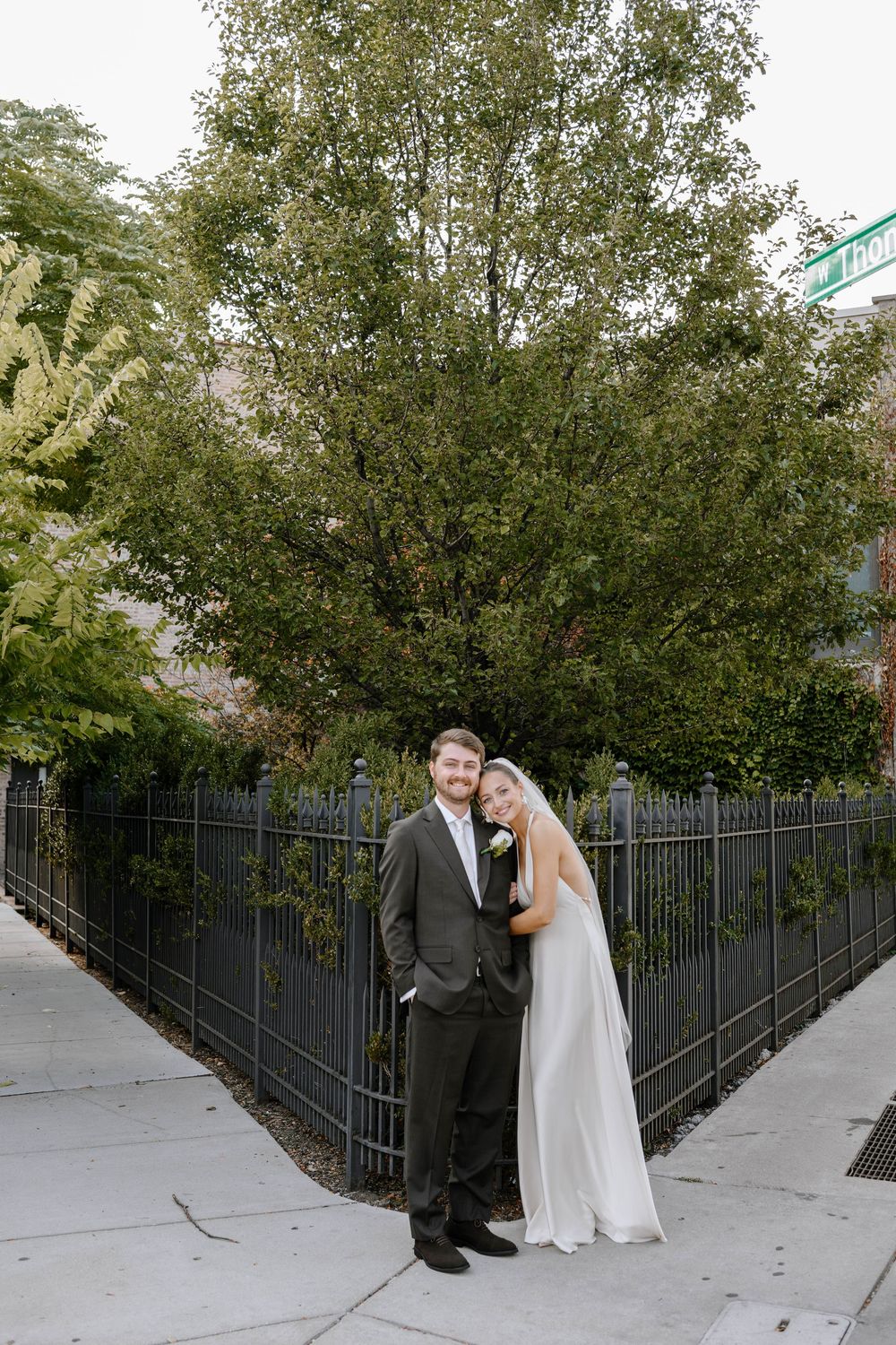 A couple in wedding attire poses together on a sidewalk with black metal fencing and trees in the background.