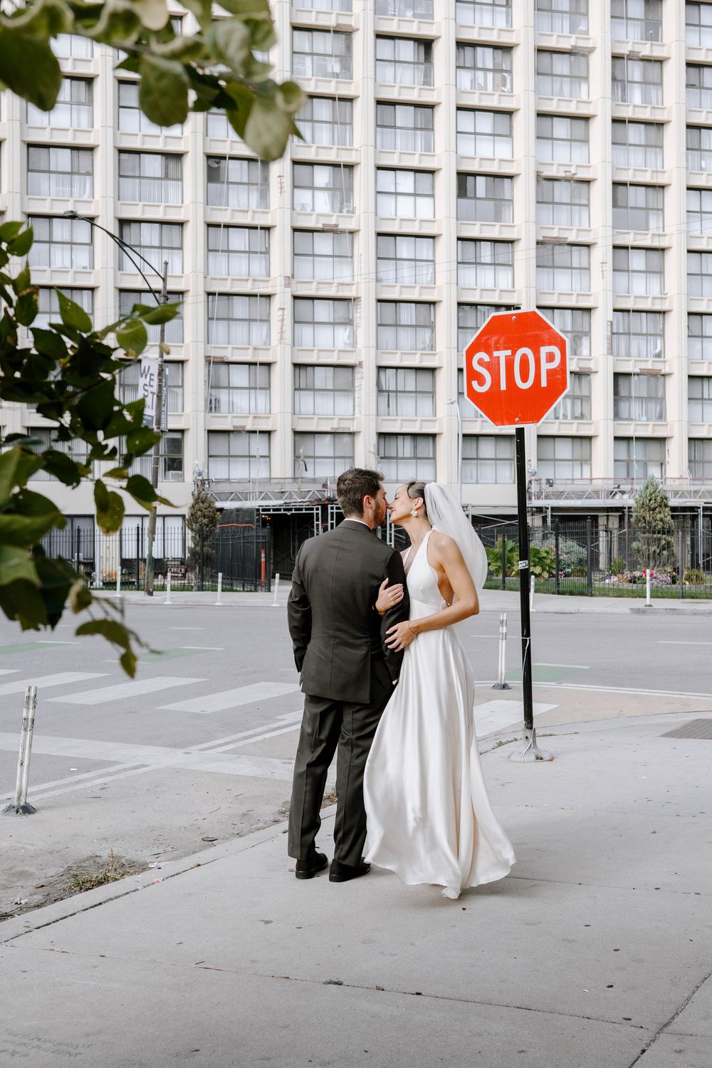 Newlyweds pose for photos on an urban sidewalk with a large building and stop sign in the background.