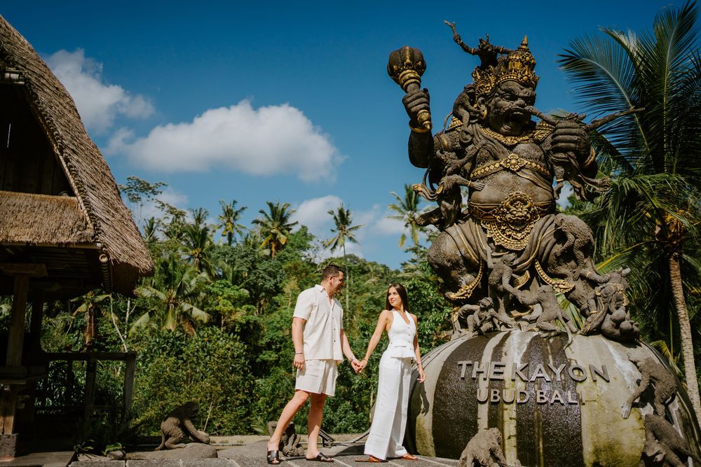 Couple poses near ornate Balinese stone statues under bright blue sky with palm trees.