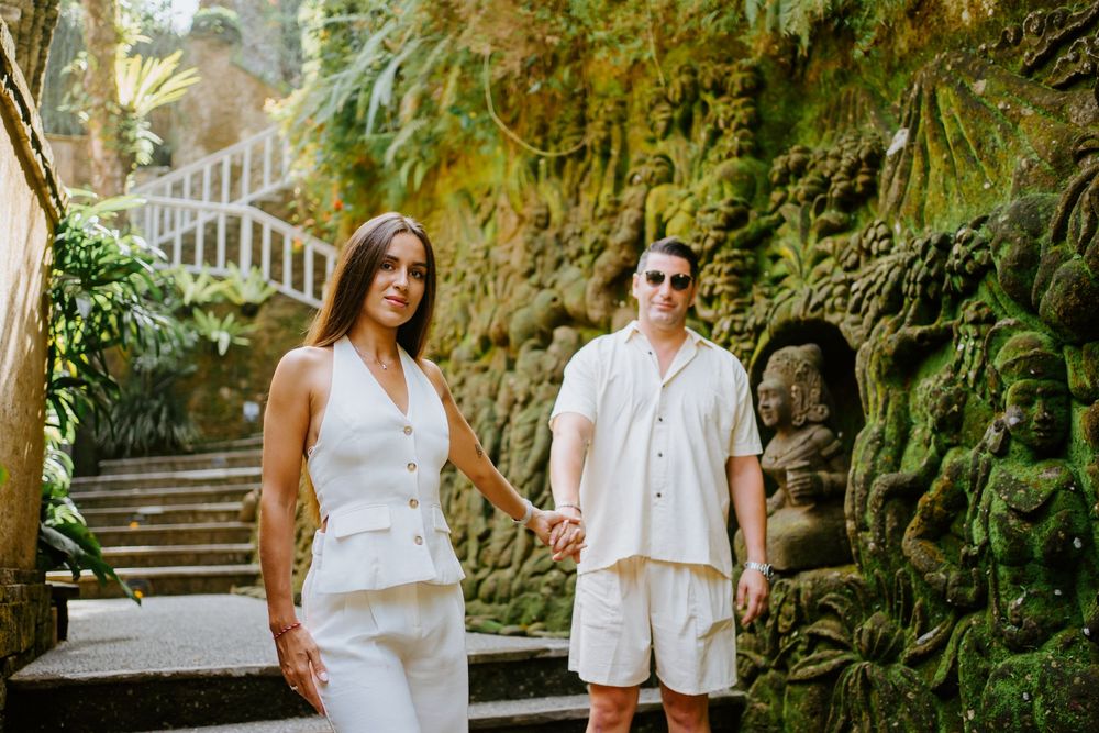Romantic couple in white outfits pose together in a lush tropical garden setting with stone steps.
