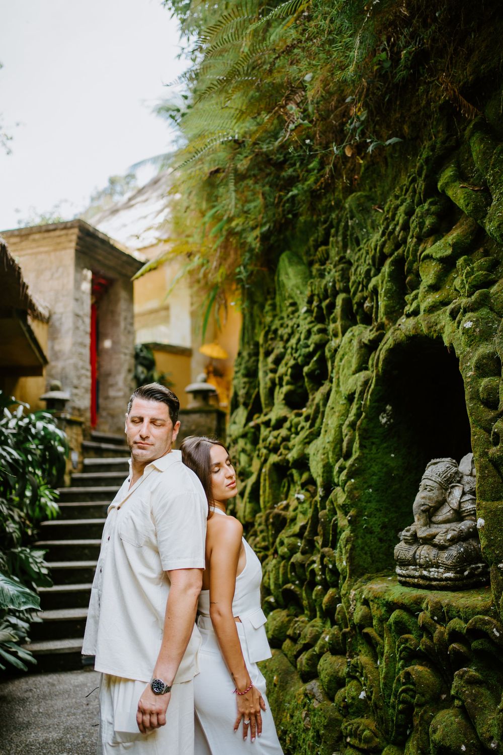 Couple shares intimate moment against mossy stone wall and tropical foliage backdrop.