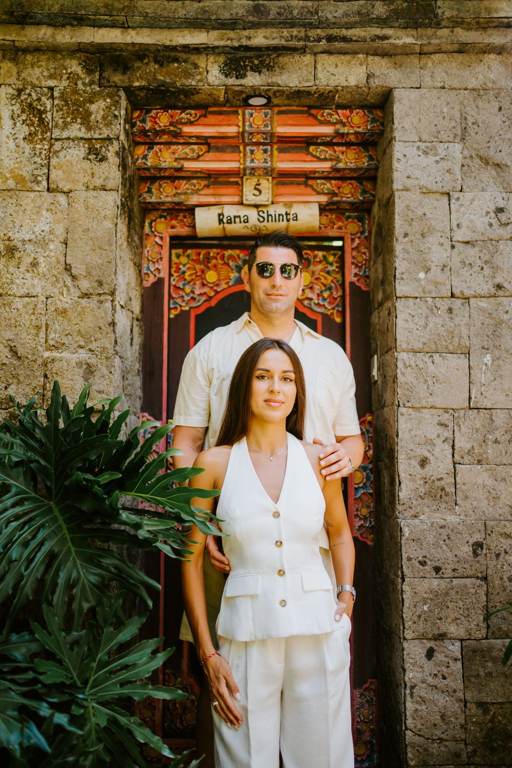 Couple in matching white outfits stand in front of ornate stone doorway with tropical plants.