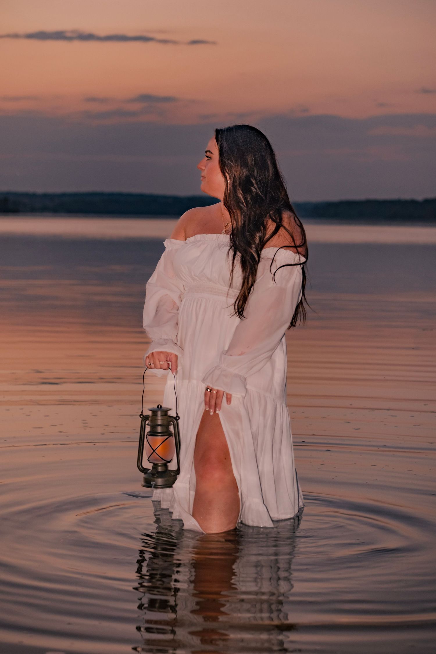A person in a white dress holds a vintage lantern while standing in calm waters during a dreamy sunset.
