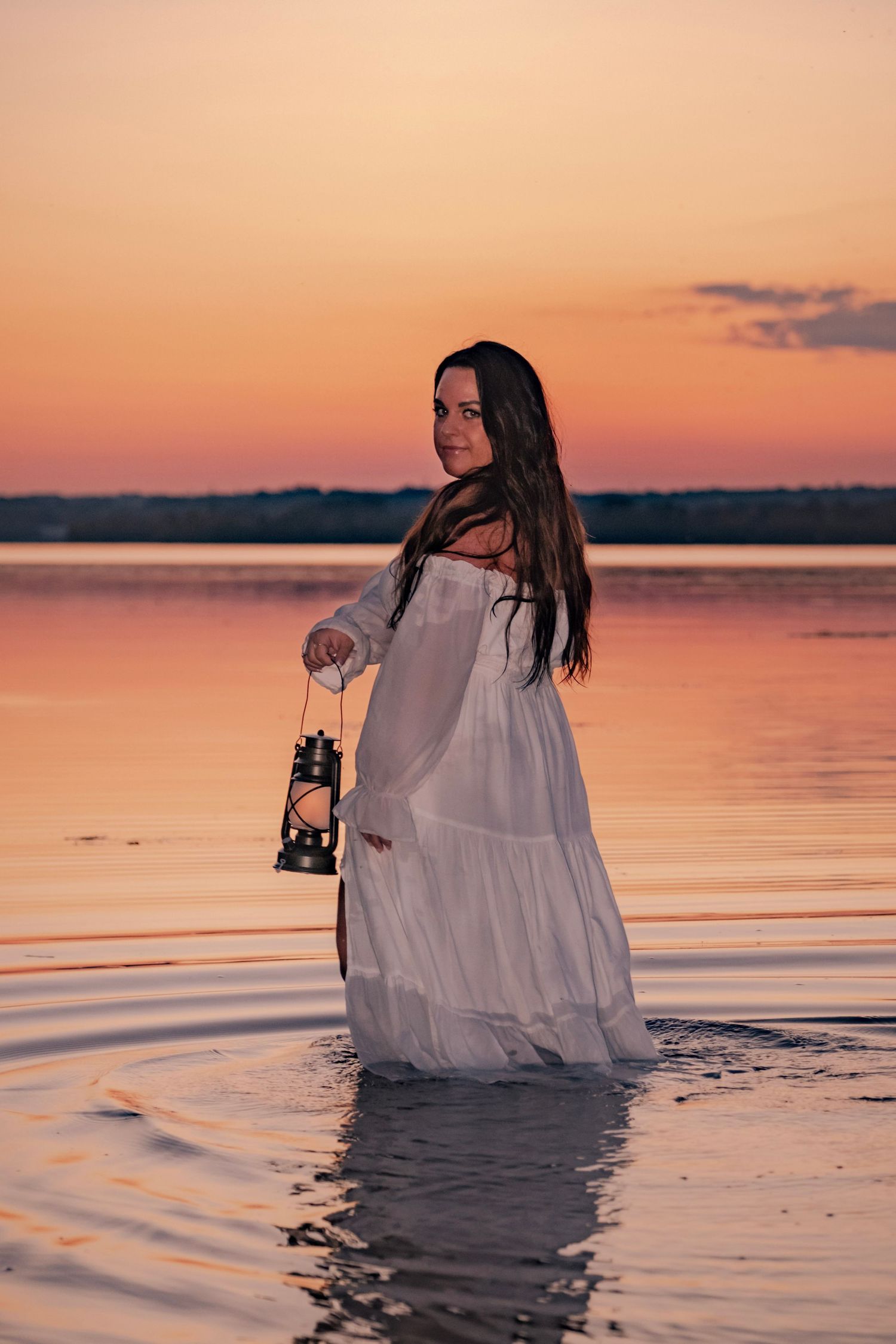 A person in a flowing white dress holds a lantern while standing in shallow water during a vibrant orange sunset.