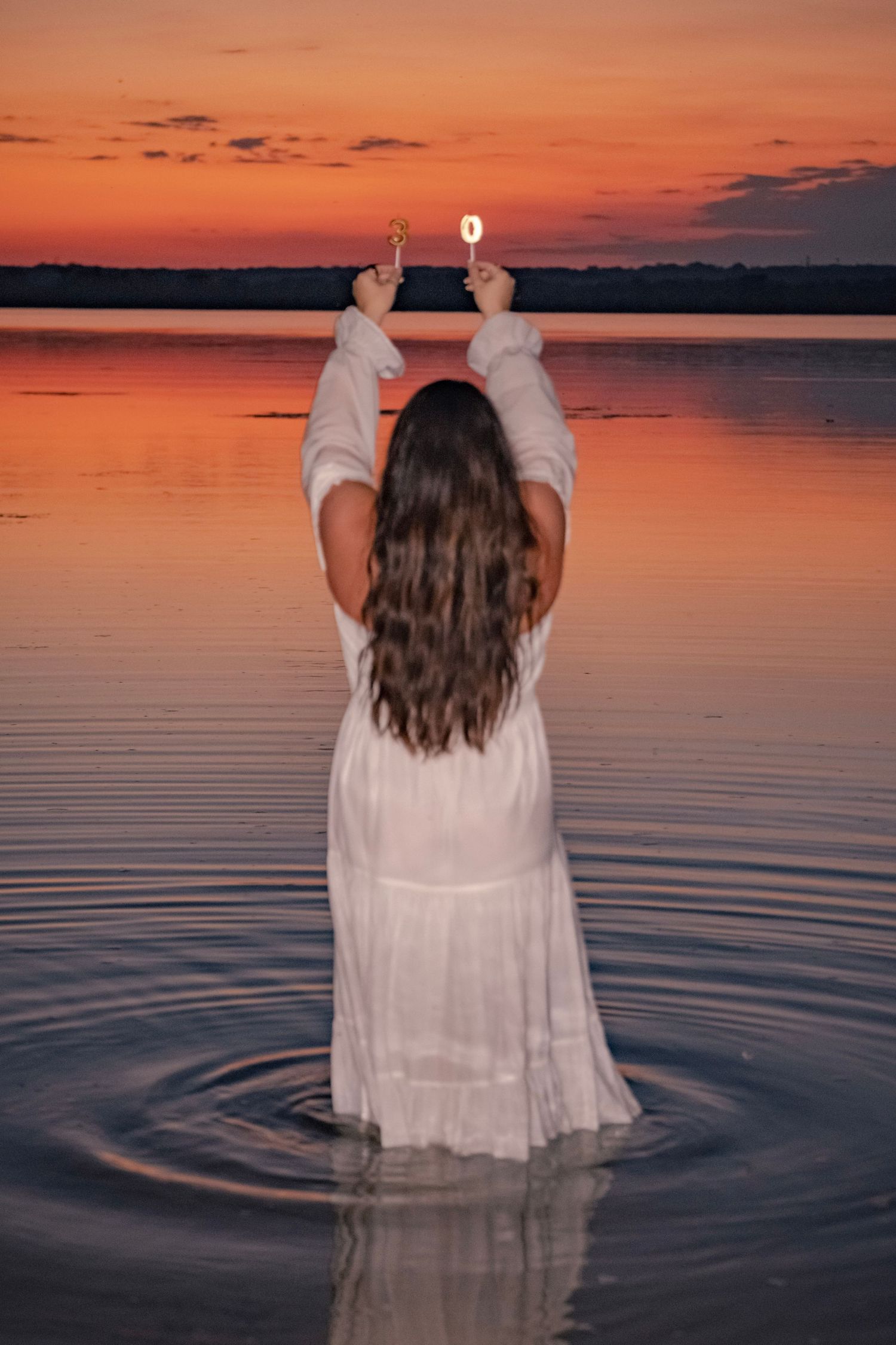 A person in a white dress stands in calm lake waters during a vibrant orange and purple sunset.