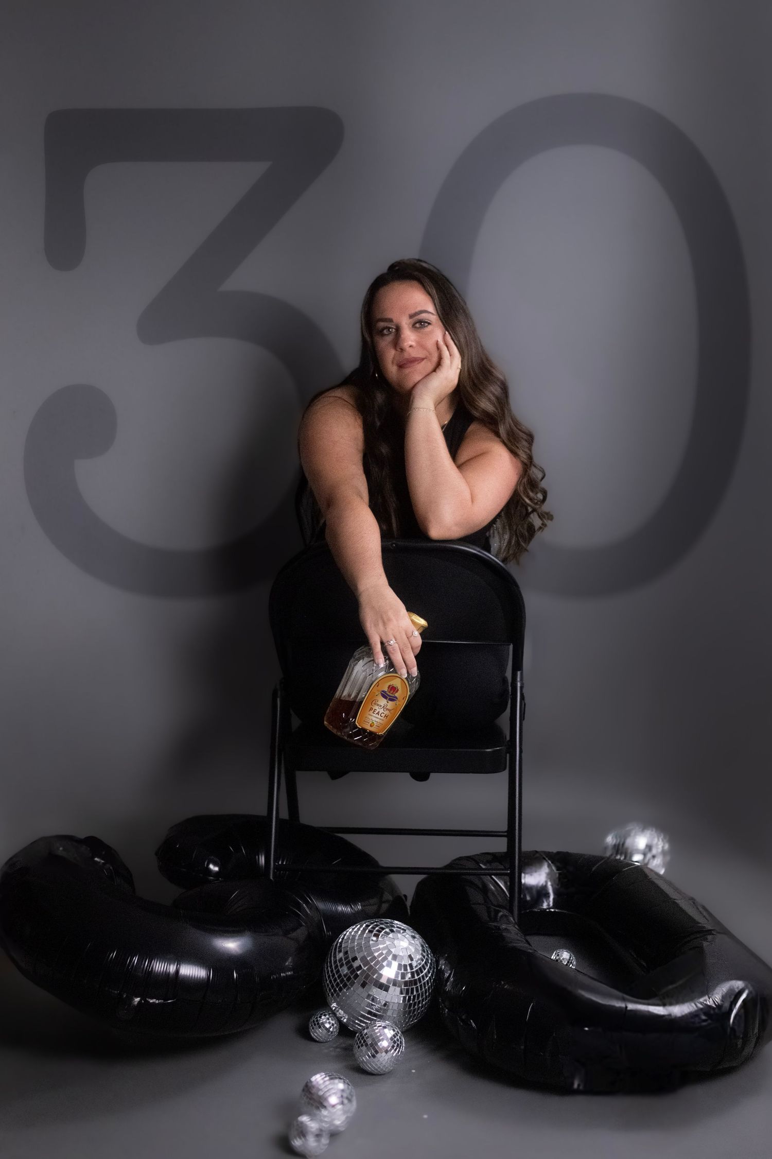 A person in black attire sits on a chair with a drink surrounded by black balloons and silver disco balls on the floor.