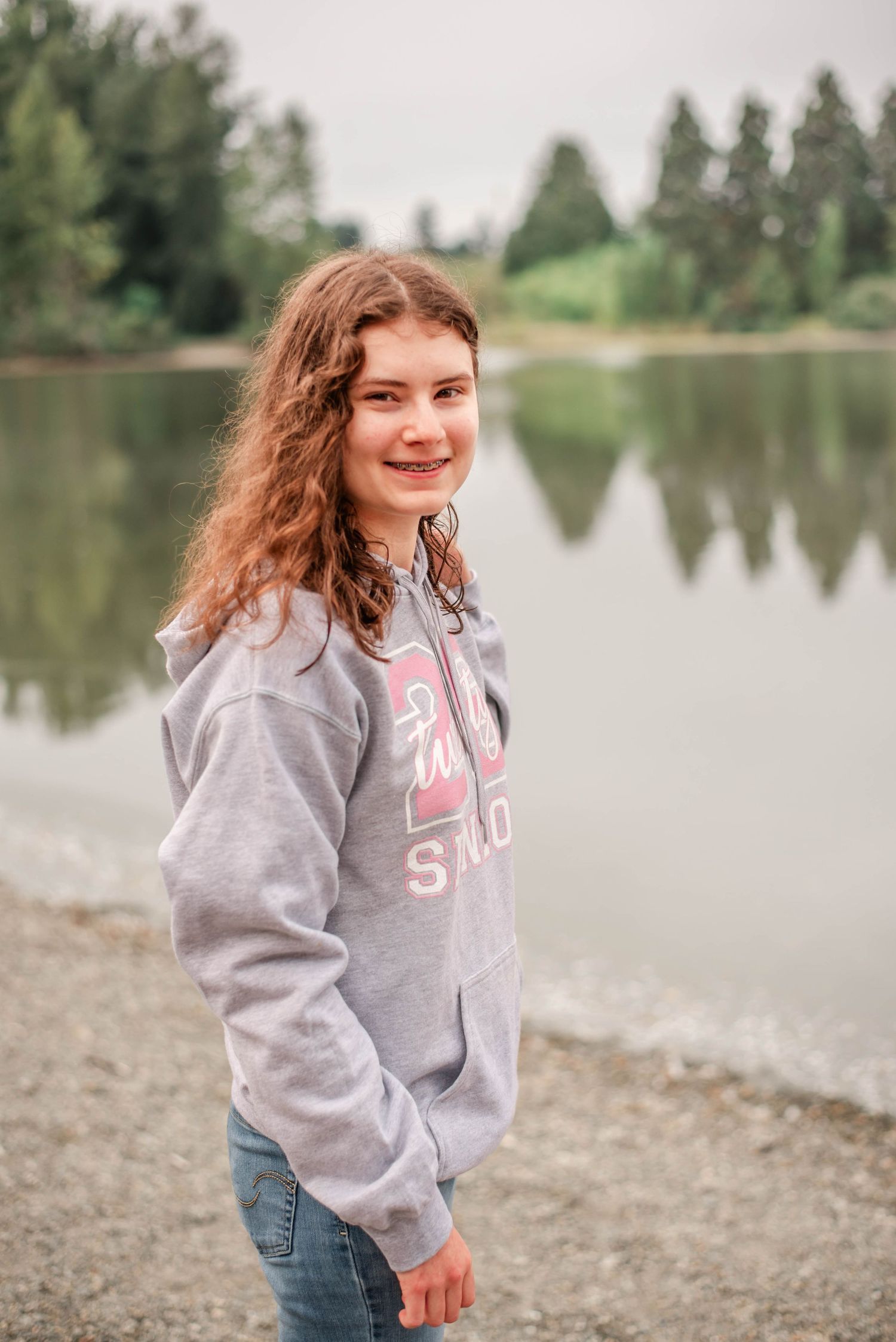A person in a gray hoodie and jeans stands by a tranquil lake with trees in the background.