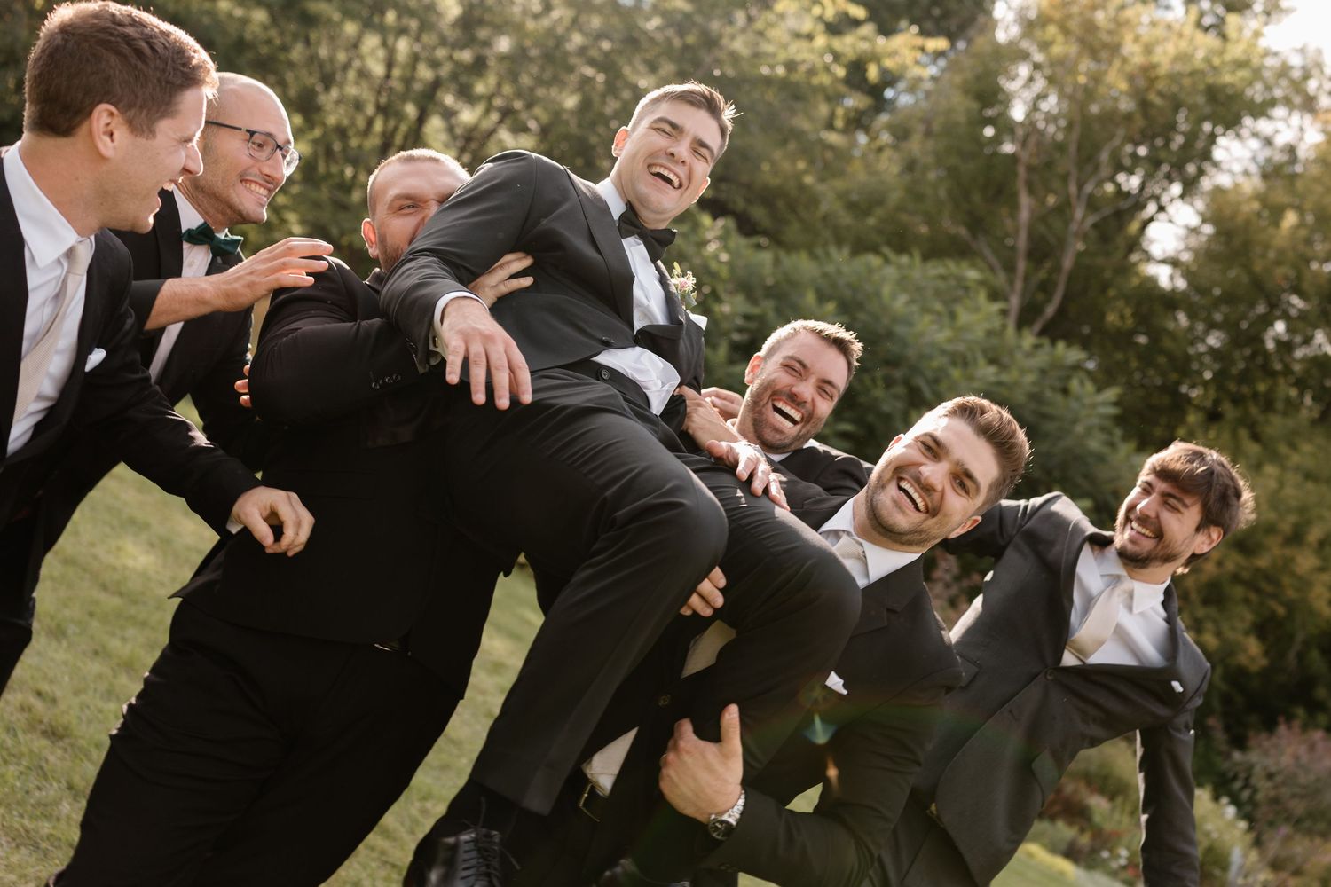 A group in formal black suits poses together outdoors while laughing and showing playful expressions during a photoshoot.