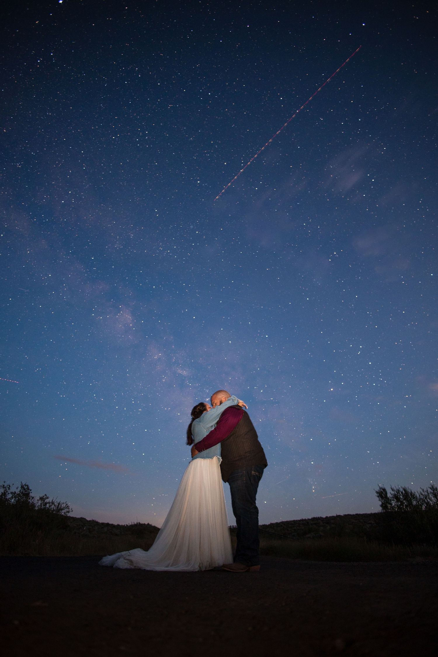 A couple poses against a starry night sky while meteor showers streak across the dark blue horizon.