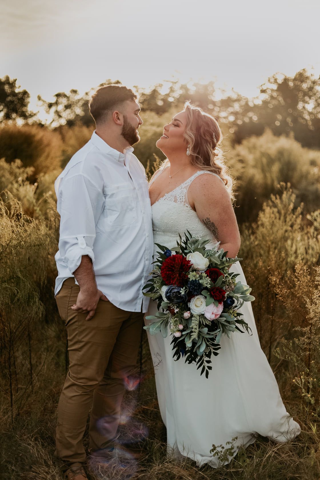 Couple shares intimate moment in golden sunlight with burgundy and white floral bouquet.