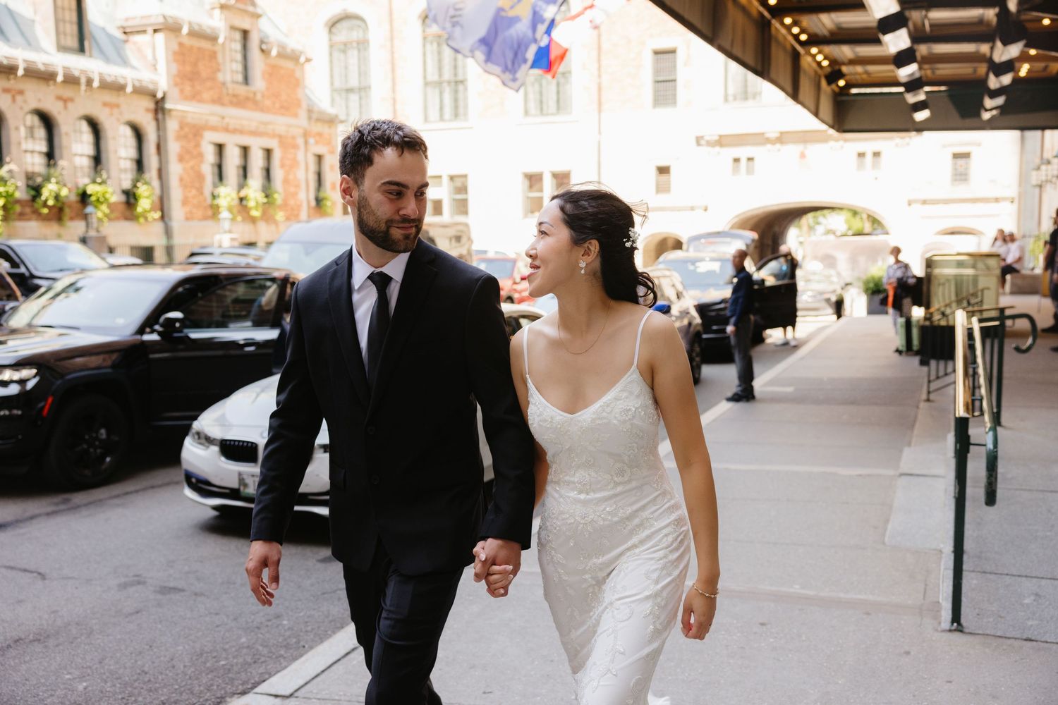 Bride in a white lace dress stands next to groom in black suit in an elegant hallway.