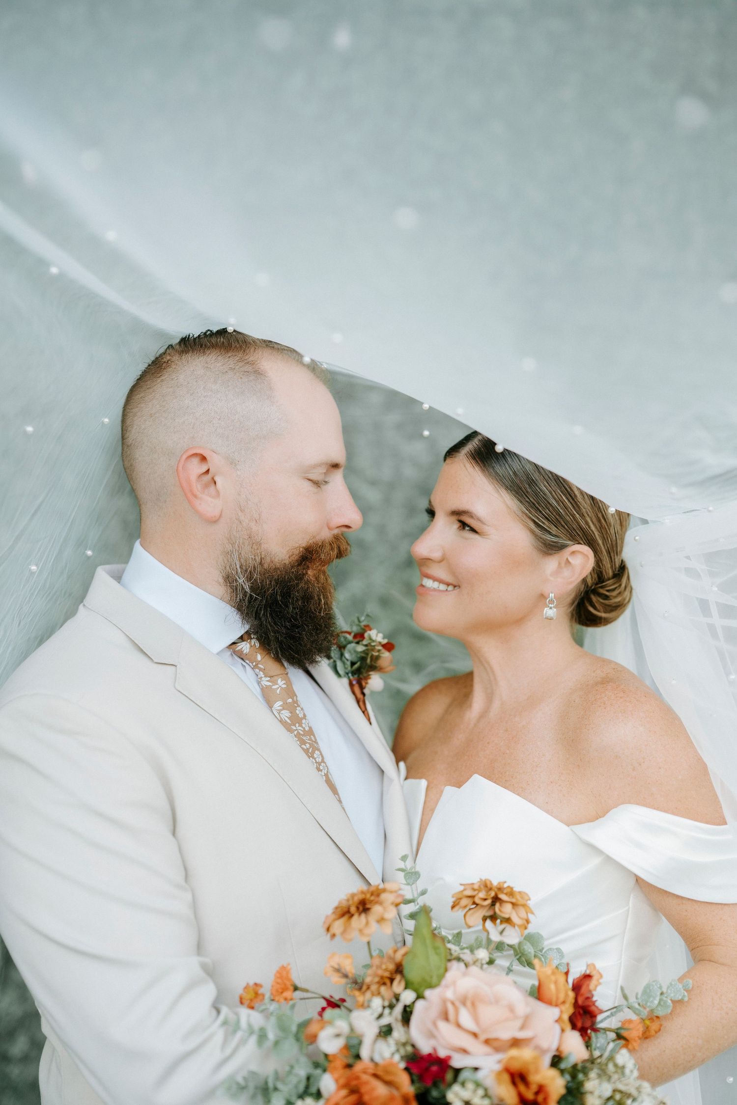 A romantic wedding portrait featuring a couple in white attire with orange and pink floral bouquet against a misty background.