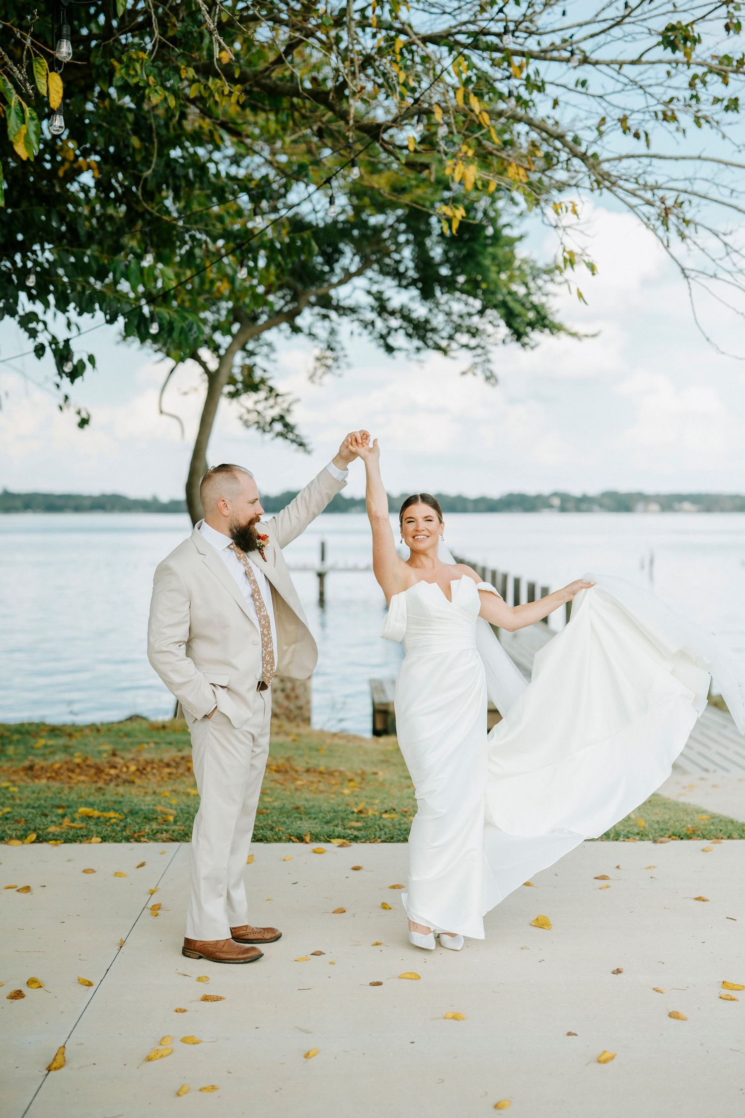 Playful wedding couple dances on a waterfront lawn with autumn leaves scattered on the ground.