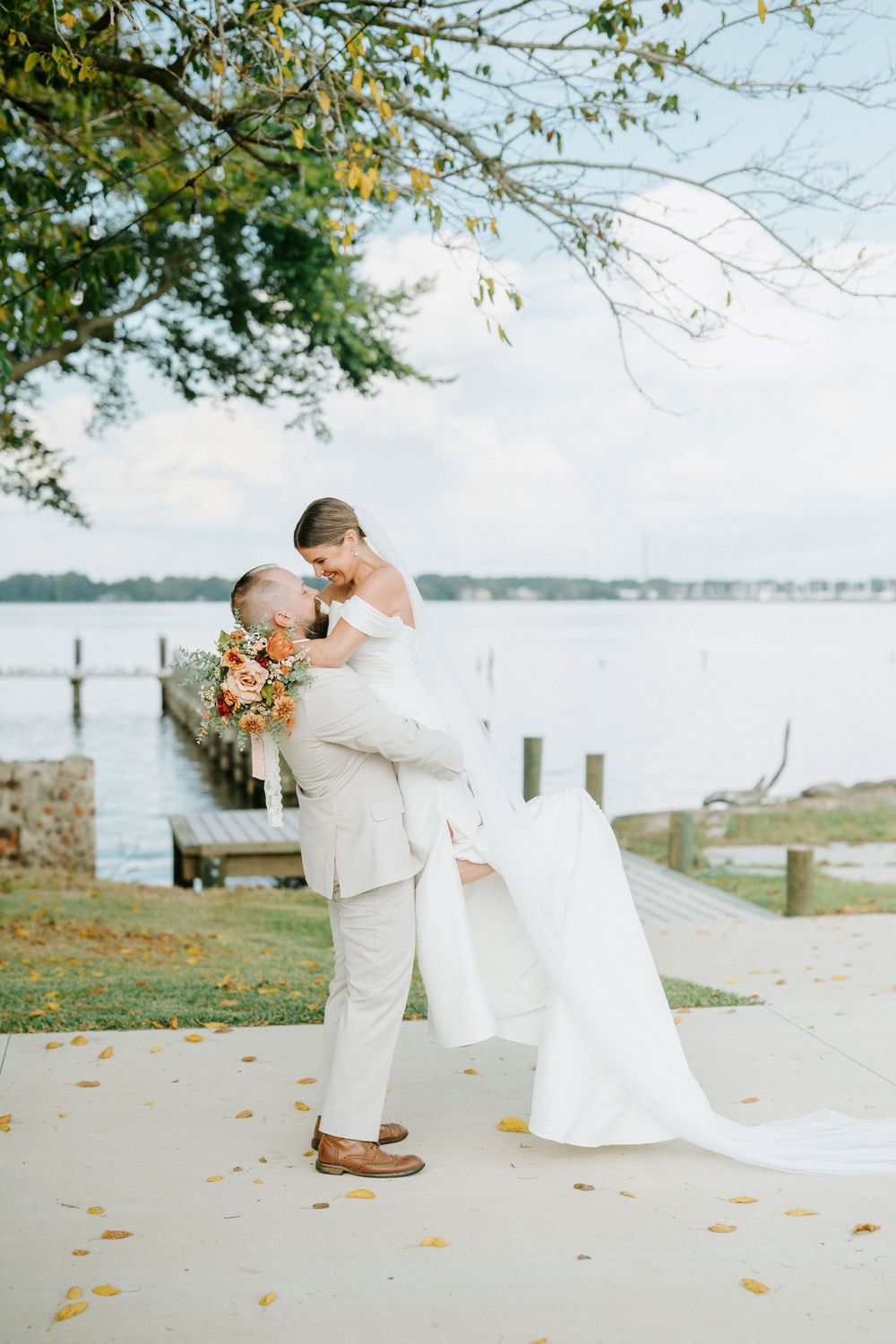 A romantic lakeside wedding moment with a couple in light-colored attire embracing near autumn trees.