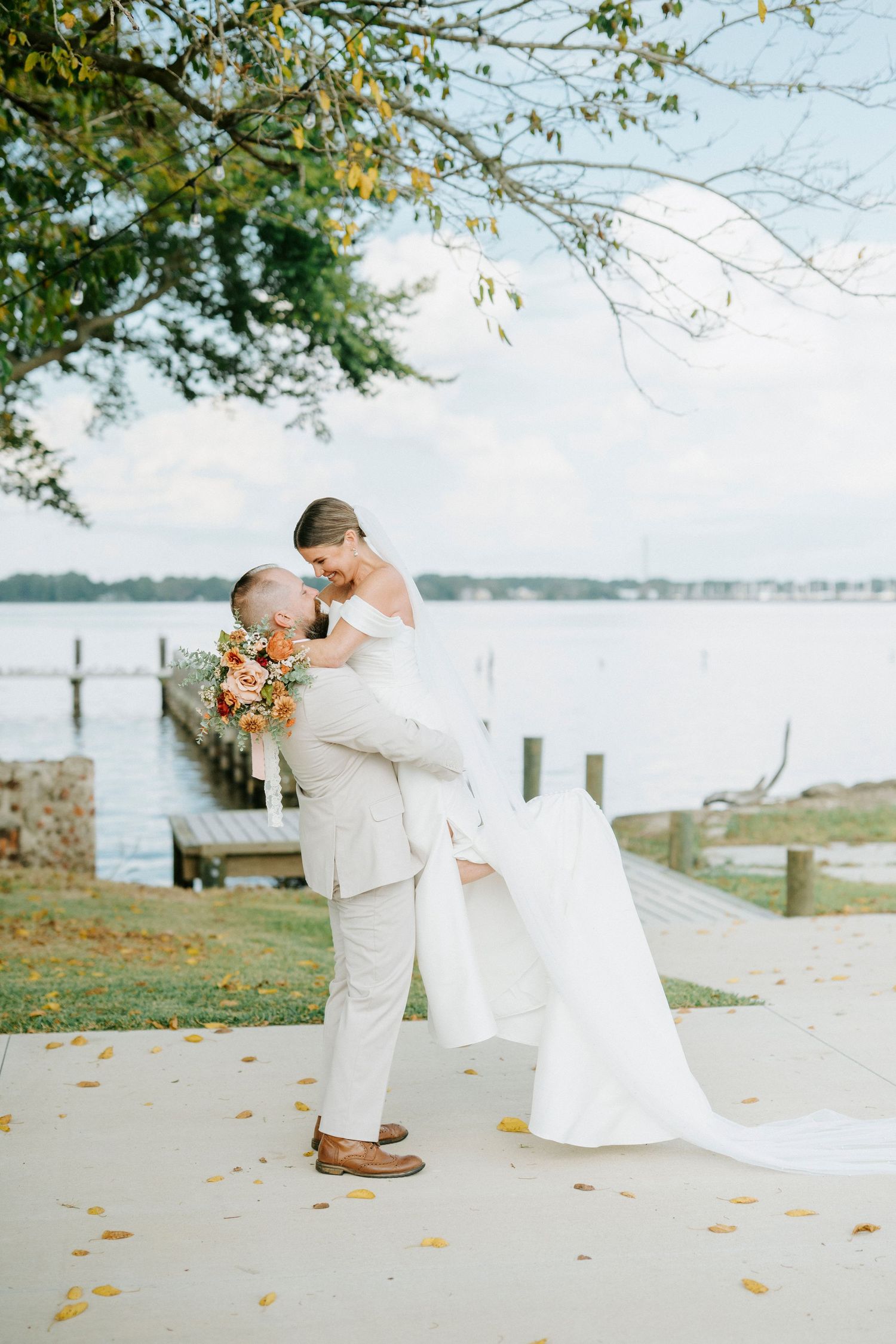 A romantic lakeside wedding moment with a couple in light-colored attire embracing near autumn trees.
