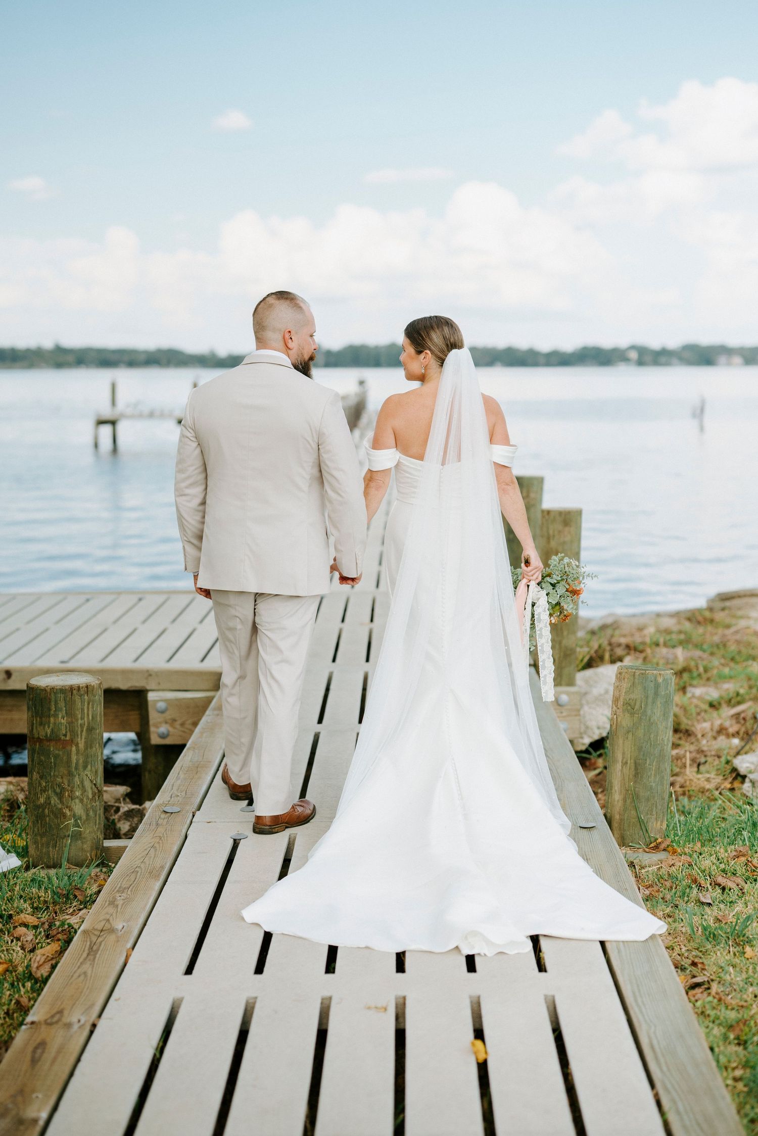 A couple in wedding attire sits together on a wooden dock overlooking a serene waterfront view.