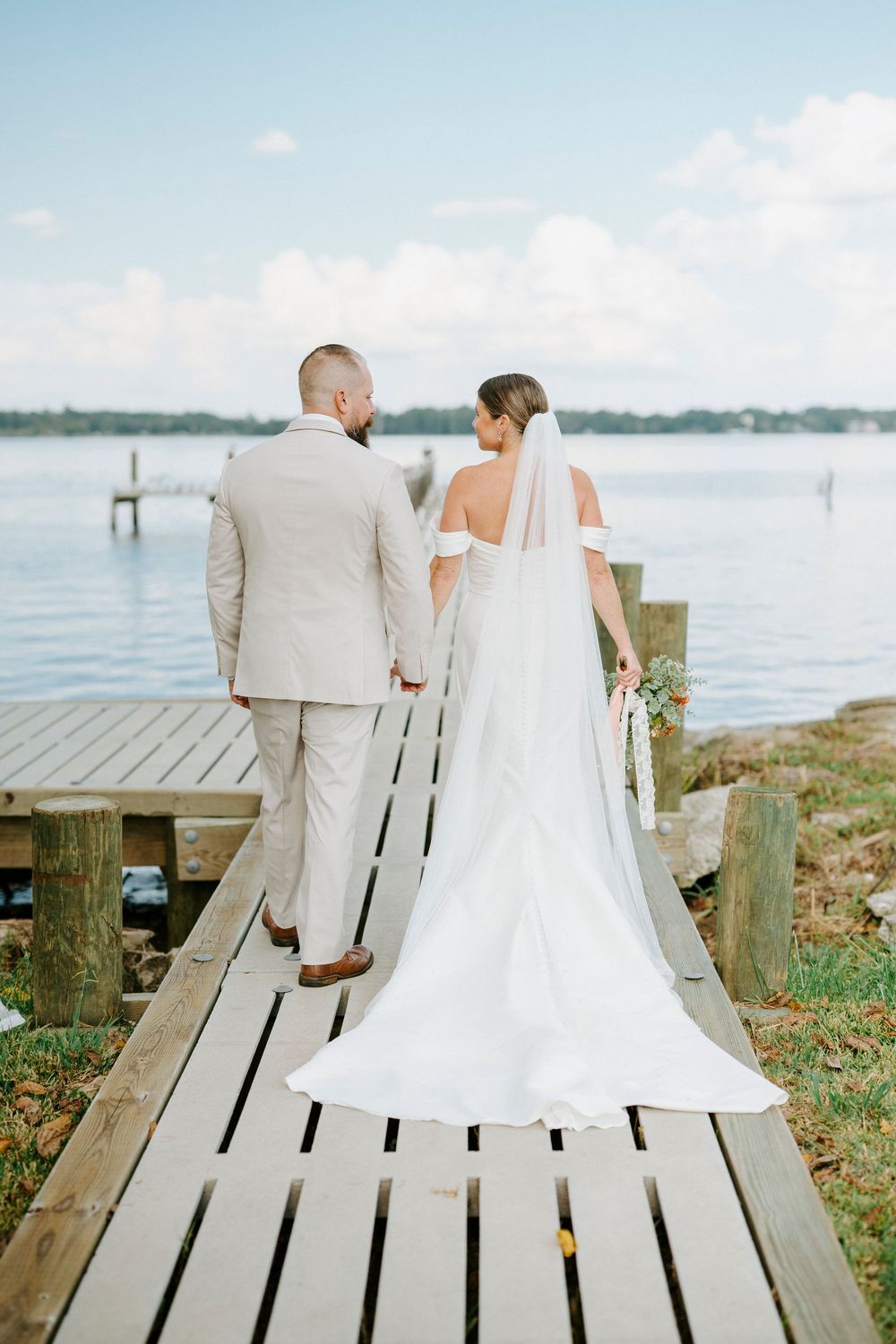 A couple in wedding attire sits together on a wooden dock overlooking a serene waterfront view.