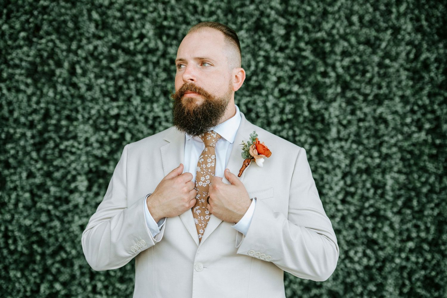 A groom in a light gray suit adjusts his bowtie against a lush green hedge backdrop.