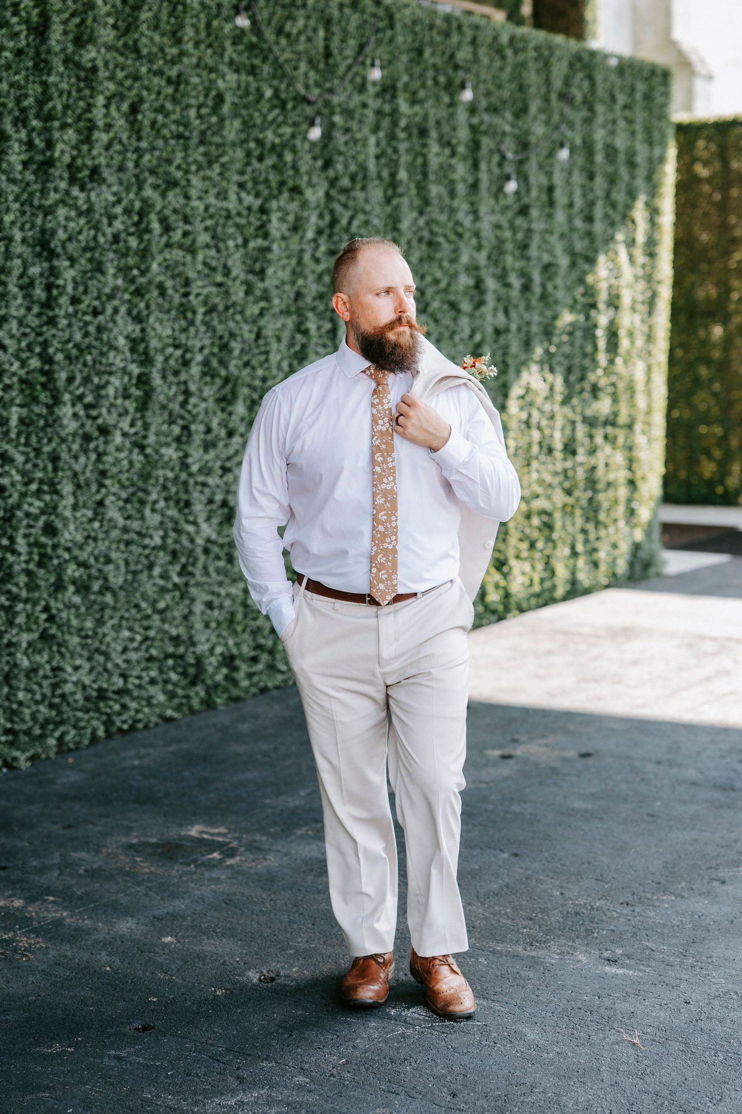 A person in a white suit with a floral tie poses against a lush green hedge wall backdrop.