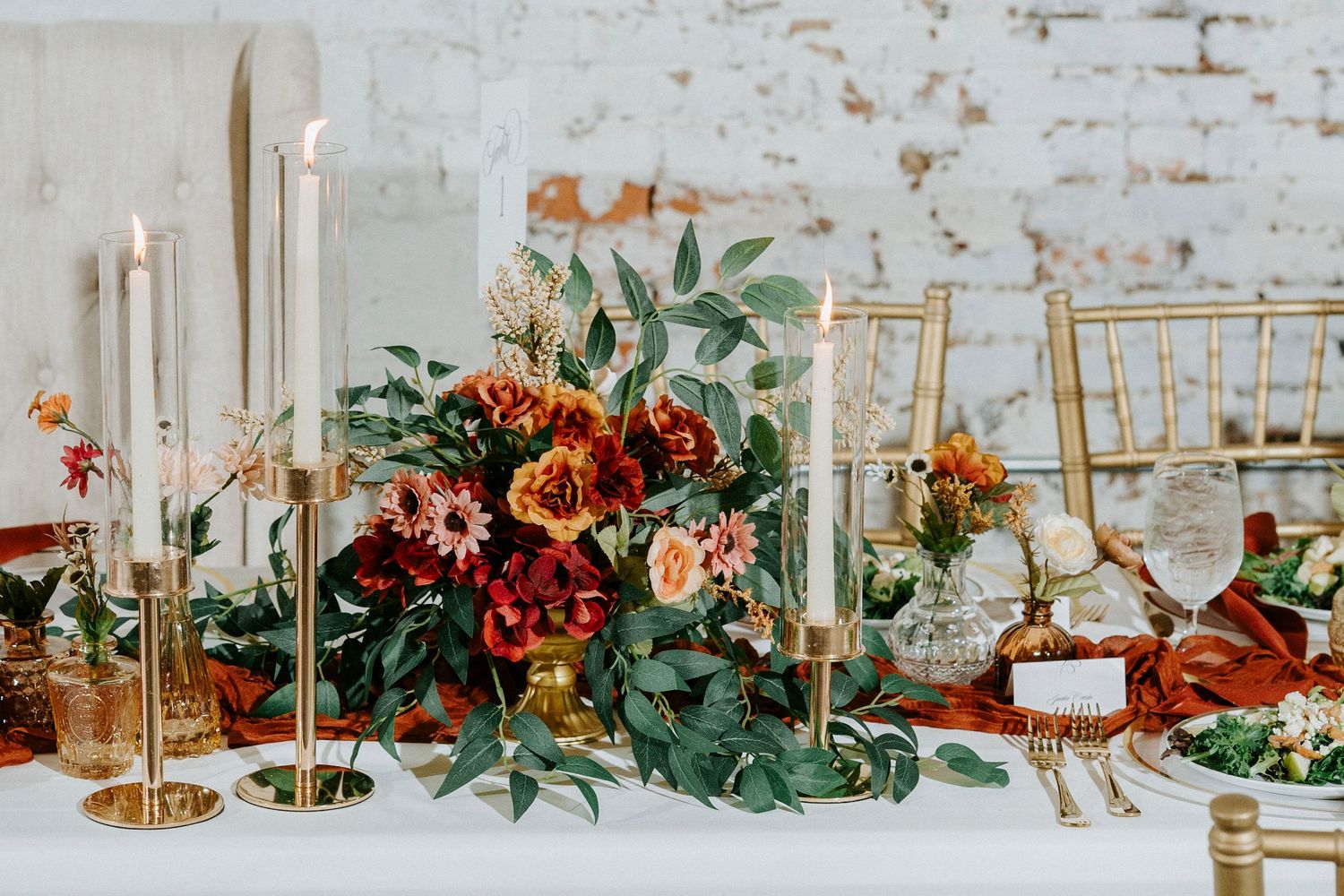 Elegant wedding table decor with greenery, gold candlesticks, and rustic brick wall backdrop in natural light.