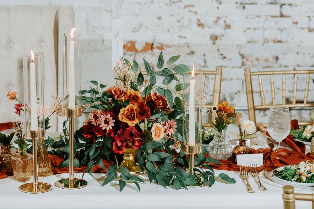 Elegant wedding table decor with greenery, gold candlesticks, and rustic brick wall backdrop in natural light.
