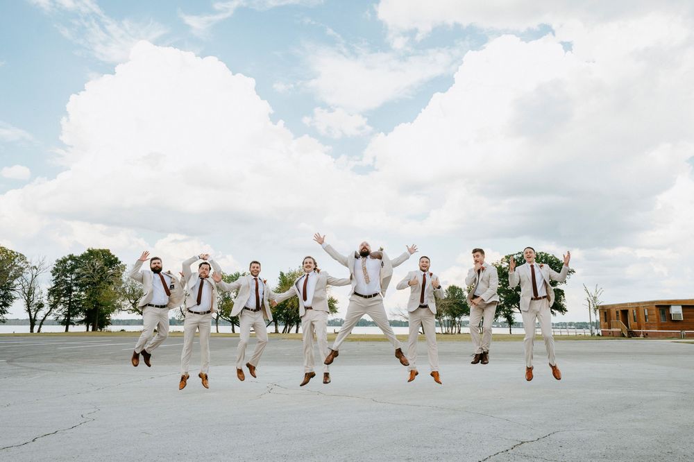 Wedding party members in white outfits jumping in synchronized motion against a cloudy sky background.
