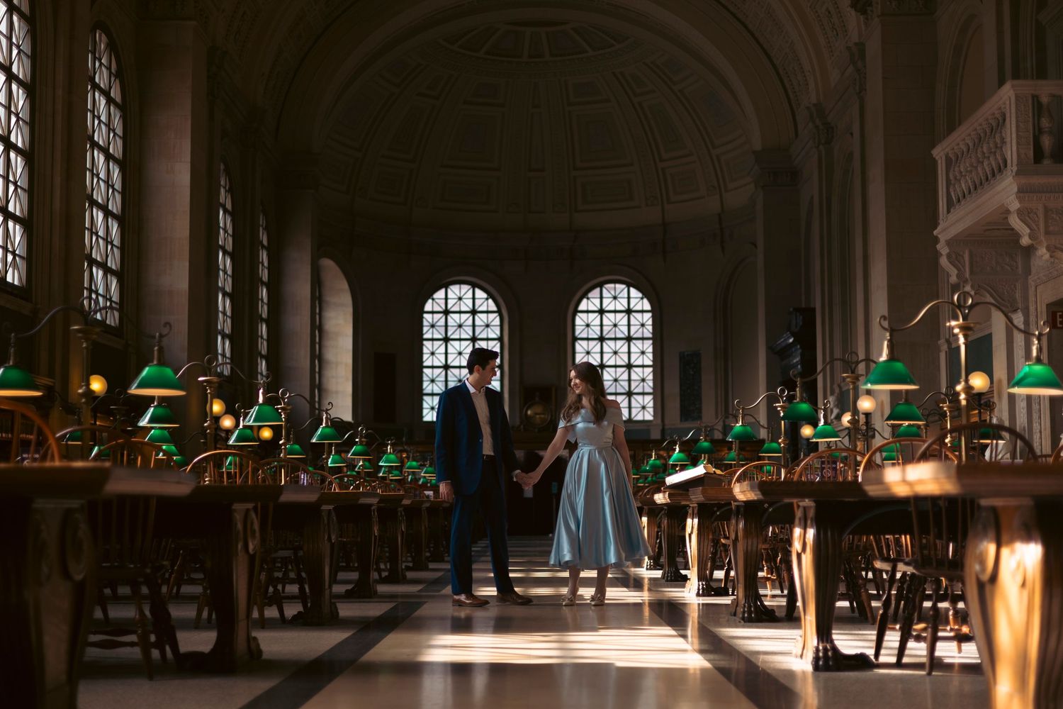 Two silhouetted figures standing in a grand library hall with green desk lamps illuminating wooden reading tables.