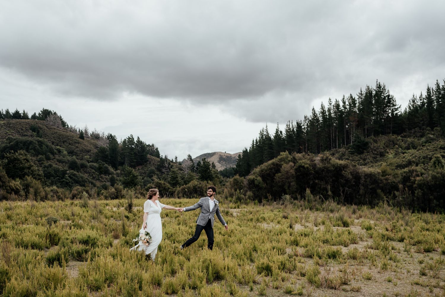 Bride and groom run across a mountain meadow with dramatic cloudy skies above during their heli-elopement in Wairarapa by August & Vine