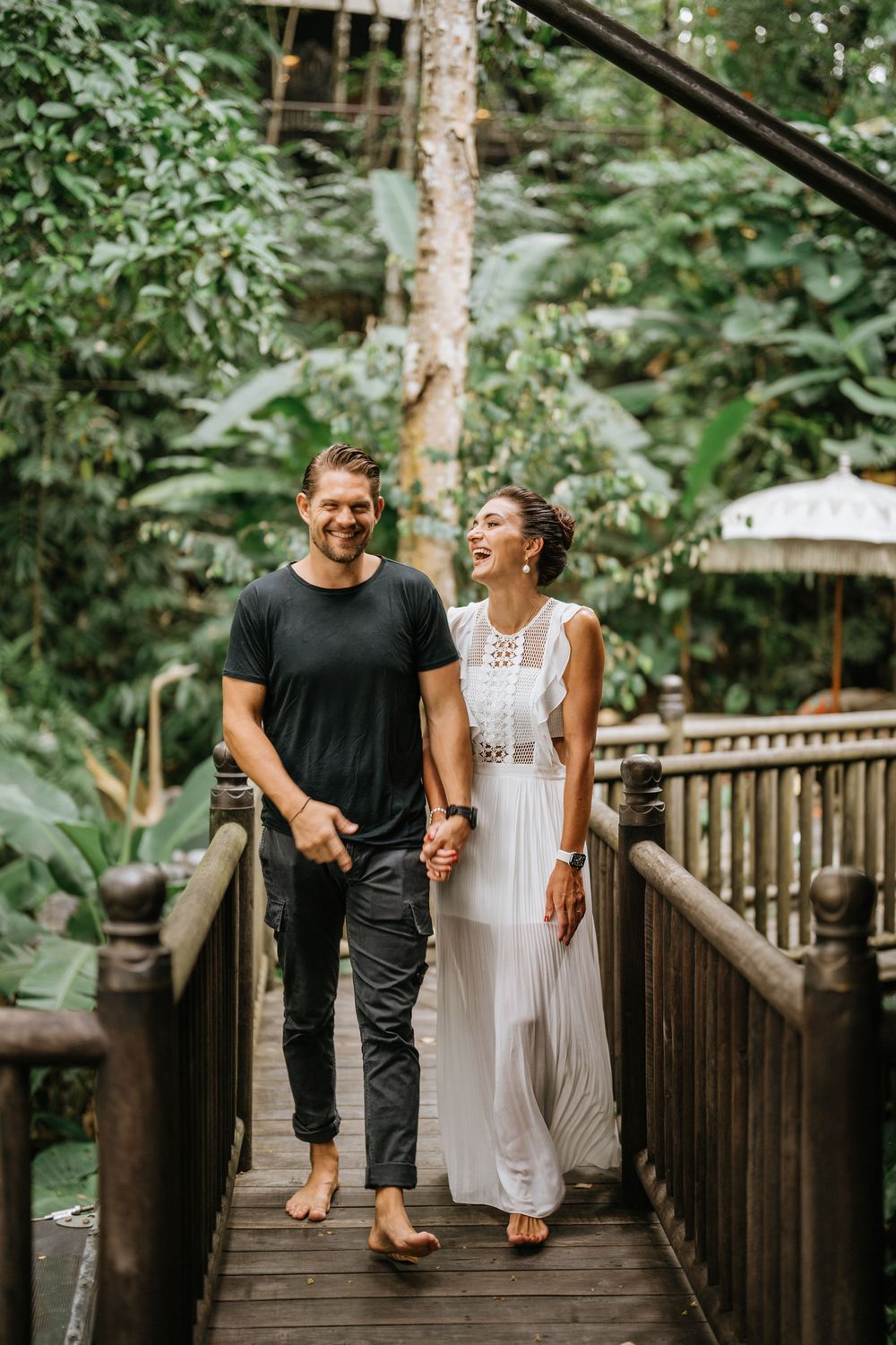 Couple walking and laughing together on wooden boardwalk surrounded by tropical trees.