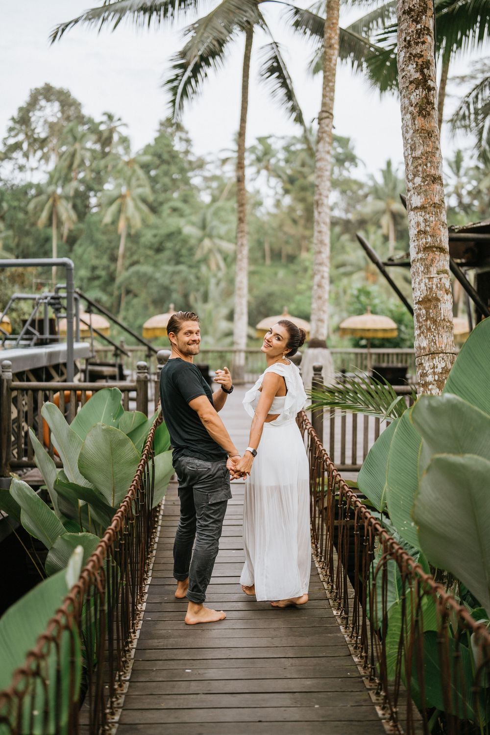 Couple sharing playful moments on a wooden walkway surrounded by tropical plants and palm trees.