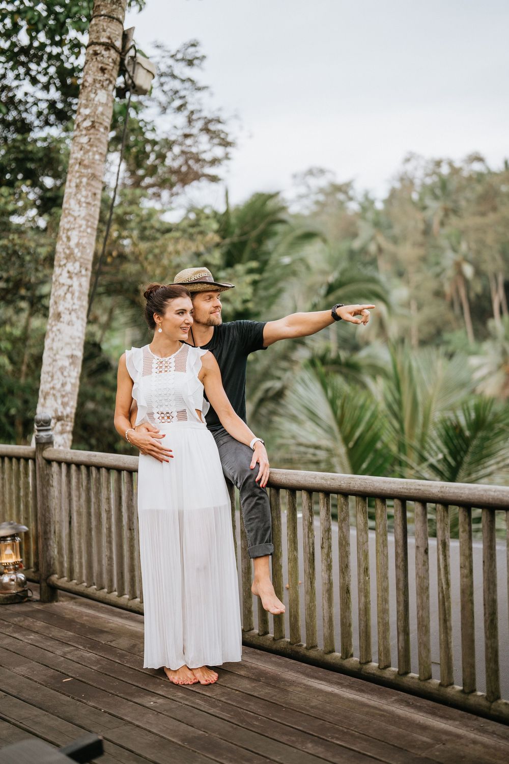 Couple standing on wooden deck overlooking tropical landscape with palm trees.