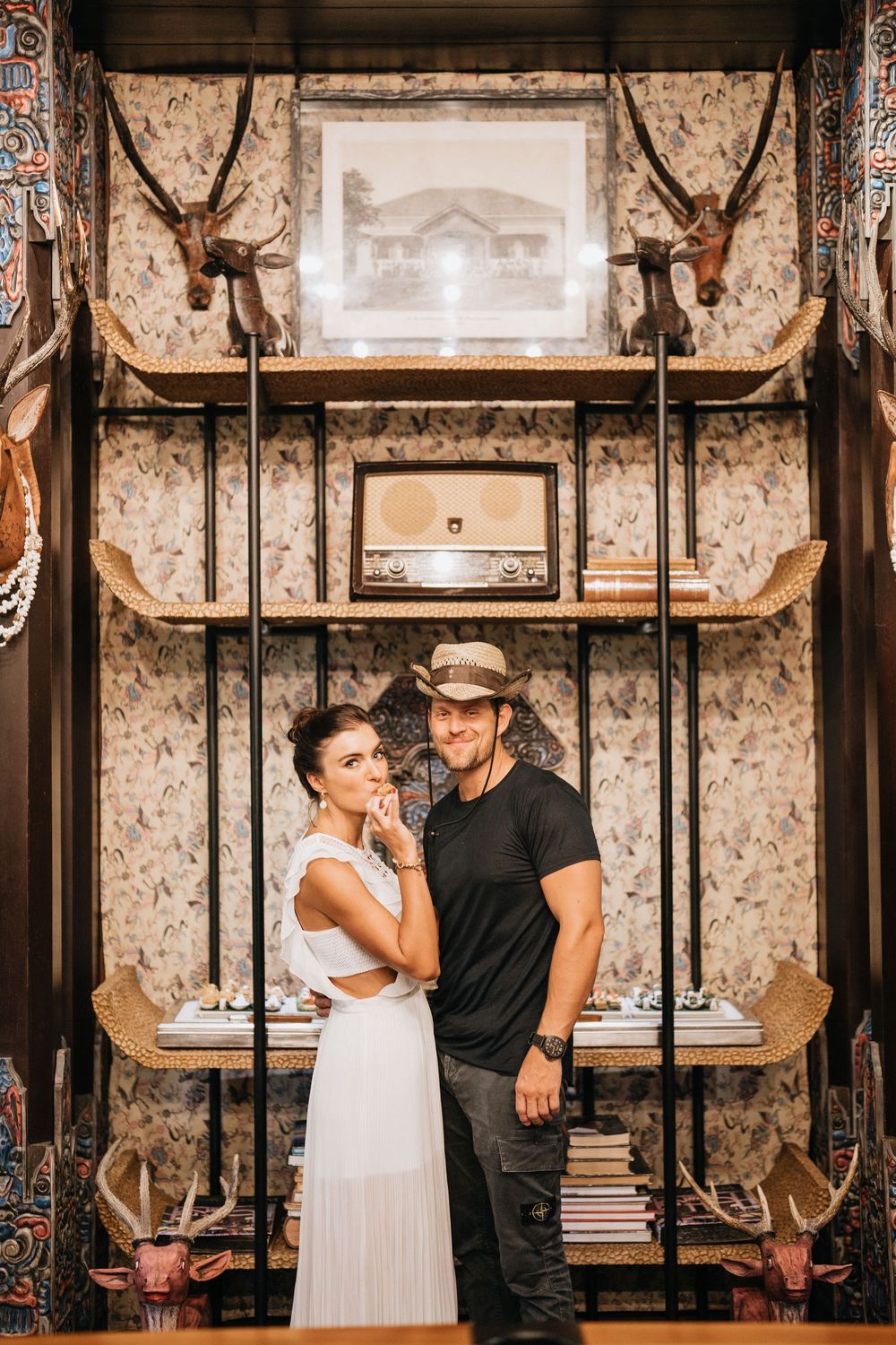 Couple posing in rustic room with antique decor and mounted antlers on textured walls.