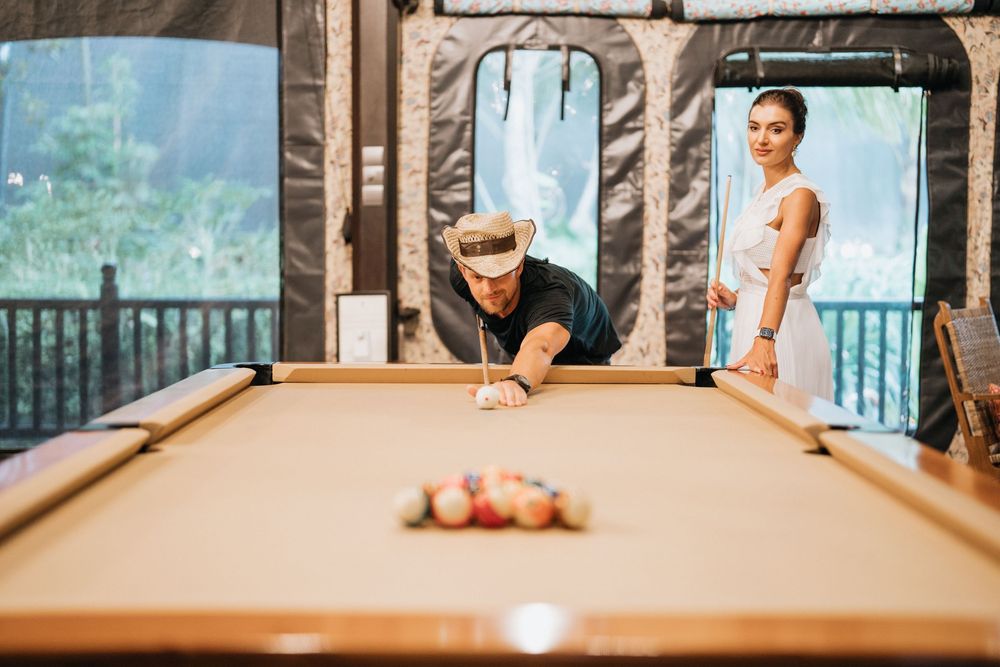 Couple playing pool in elegant room with large windows overlooking tropical scenery.