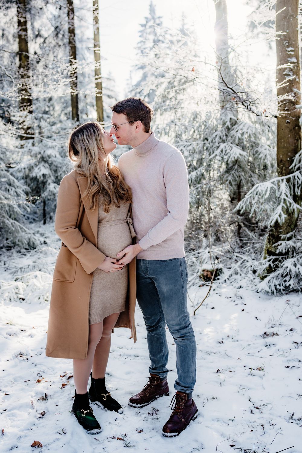 A couple shares a tender moment in a snowy forest setting wearing winter coats and boots.